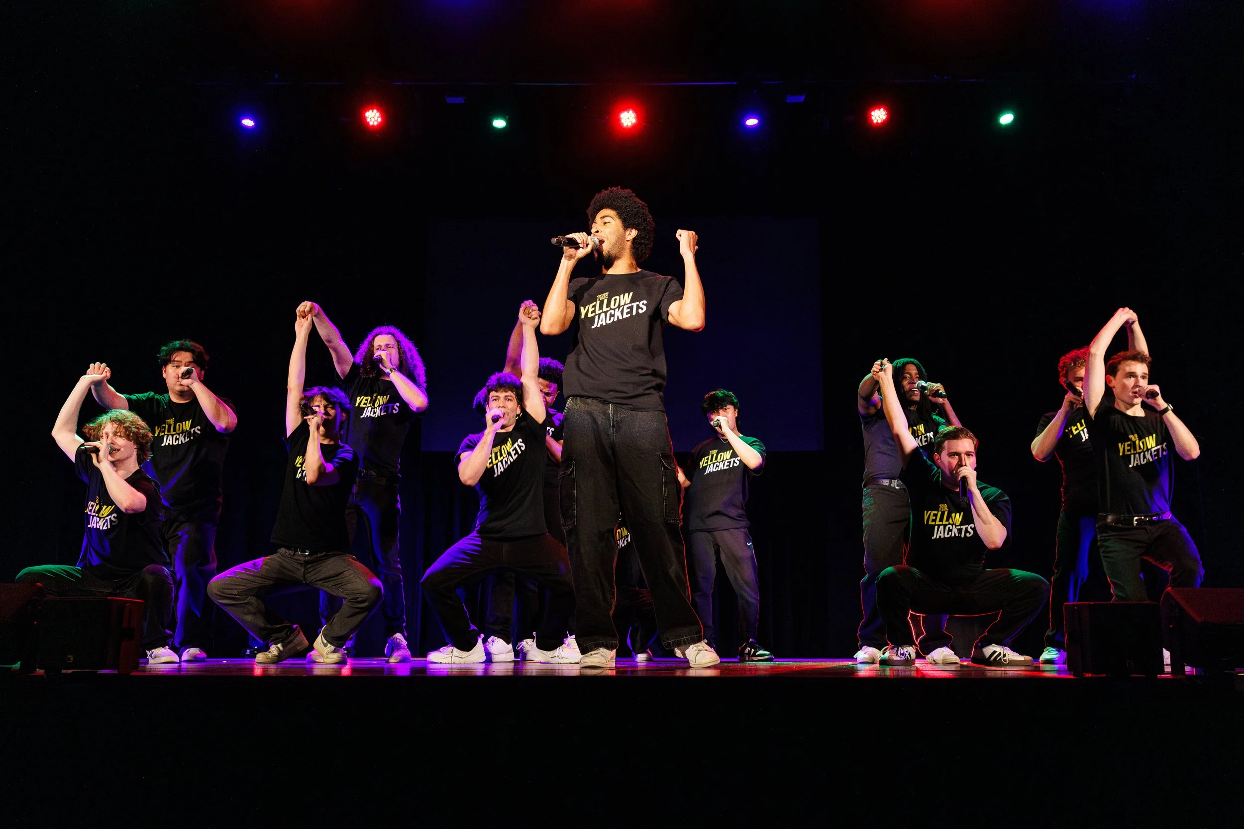 A group of young people on a stage, performing with microphones, some raising fists, wearing matching black shirts with yellow text that reads 'The Yellow Jackets,' with colorful stage lights overhead.