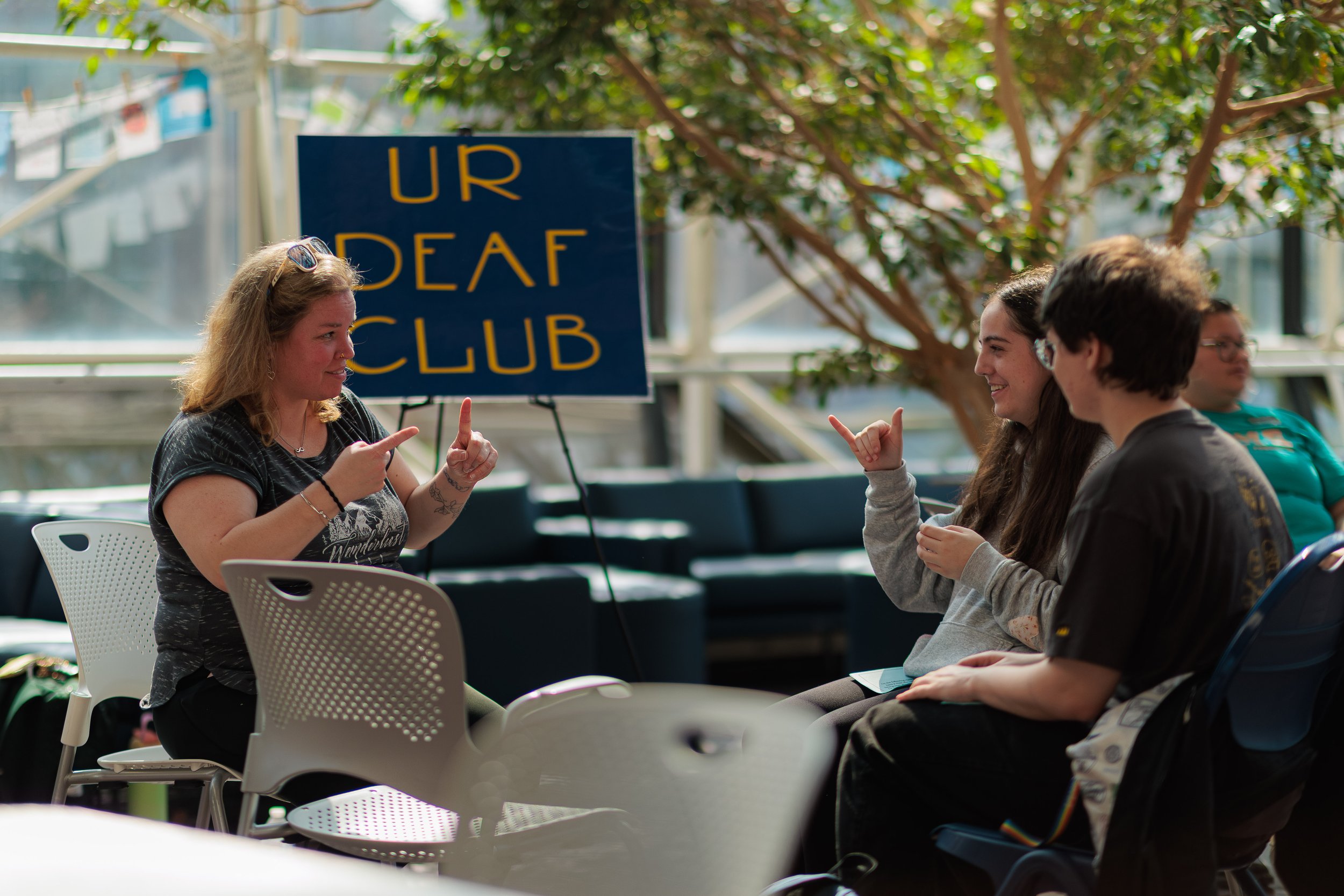 A group of four people engaged in a discussion, sitting outdoors near a sign that says 'UR DEAF CLUB'. One woman is talking and pointing with her finger, while the others are listening and smiling. There are trees and a glass building in the backgrou