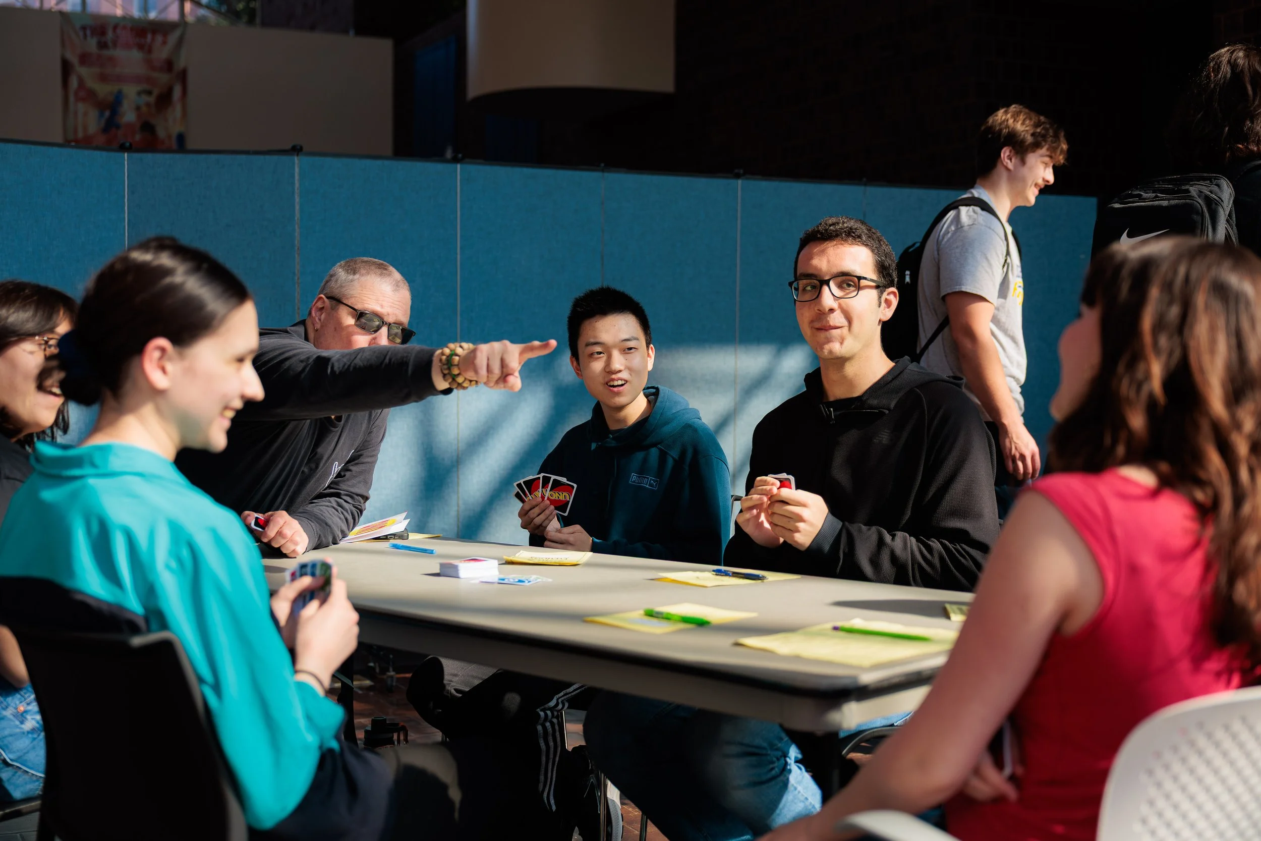 Group of young people and adult playing cards around a table indoors, engaged and smiling, with a blue wall in the background.