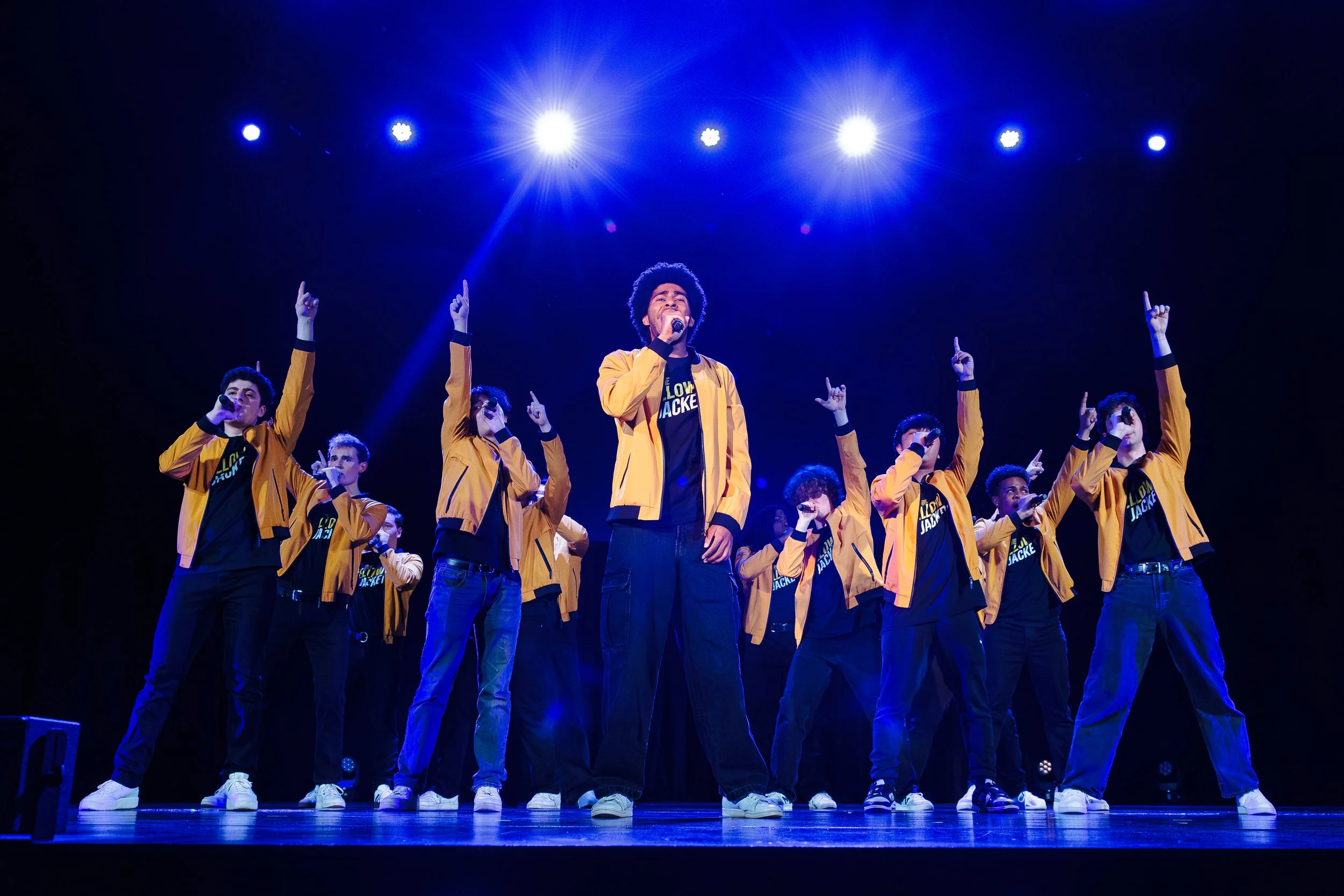 Group of performers wearing yellow jackets and black pants on stage under blue lights, singing and raising their hands.