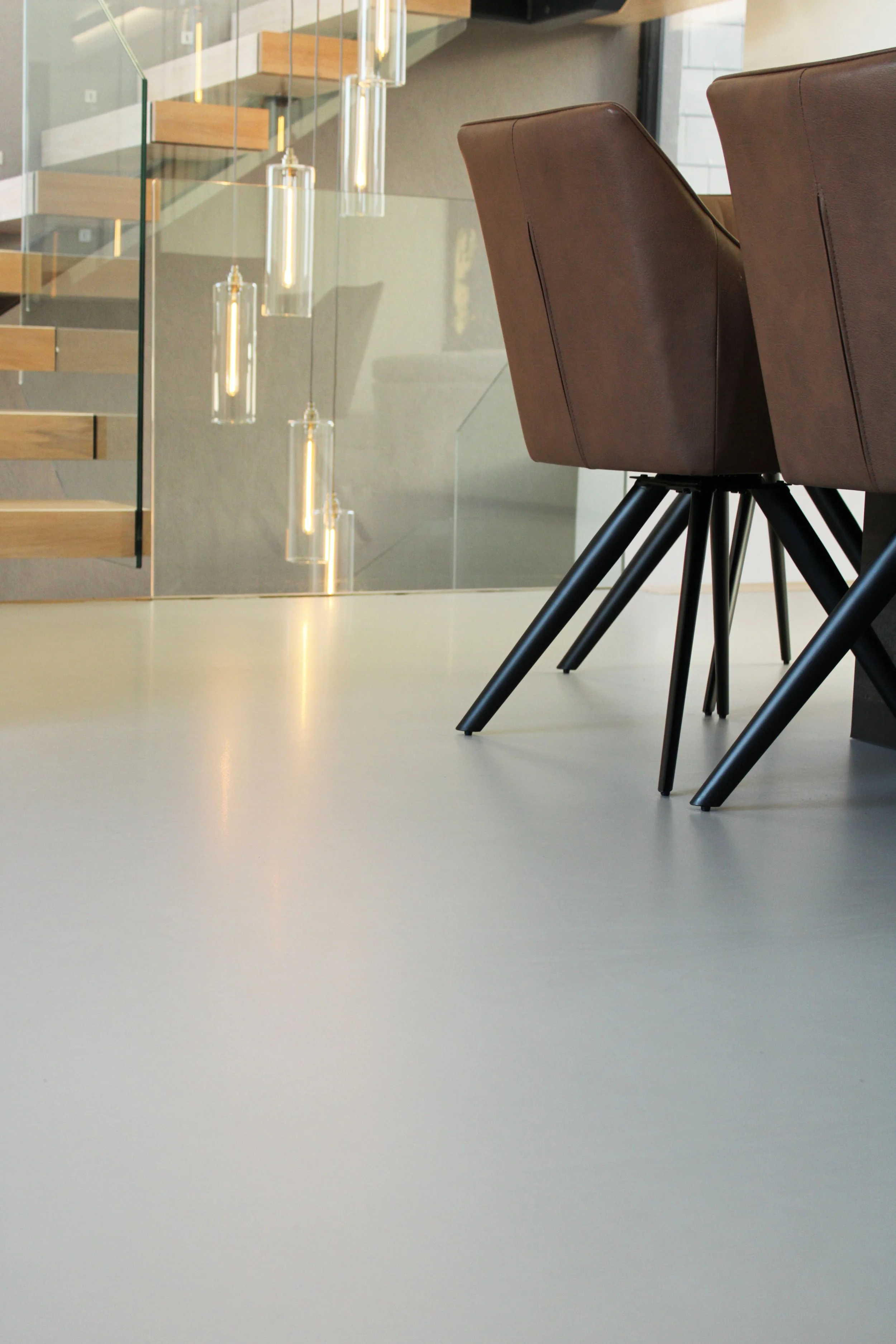 A modern interior space with two brown leather chairs with black metal legs, a glass staircase with wooden steps, and hanging pendant lights reflected on the glass.