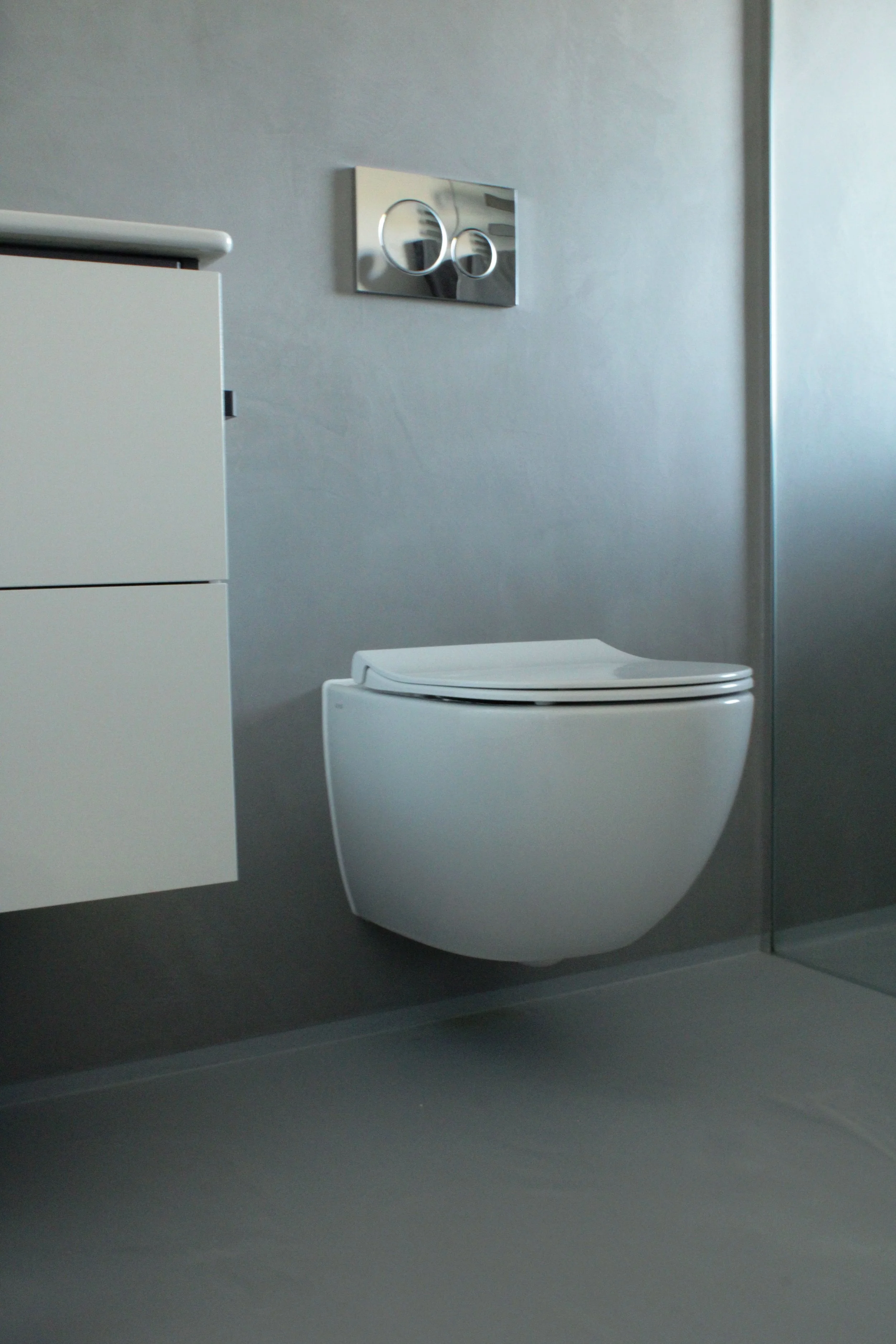 Modern bathroom with a white toilet, gray walls, and a white cabinet on the left. A metallic flush plate with two circular buttons is mounted on the wall above the toilet.