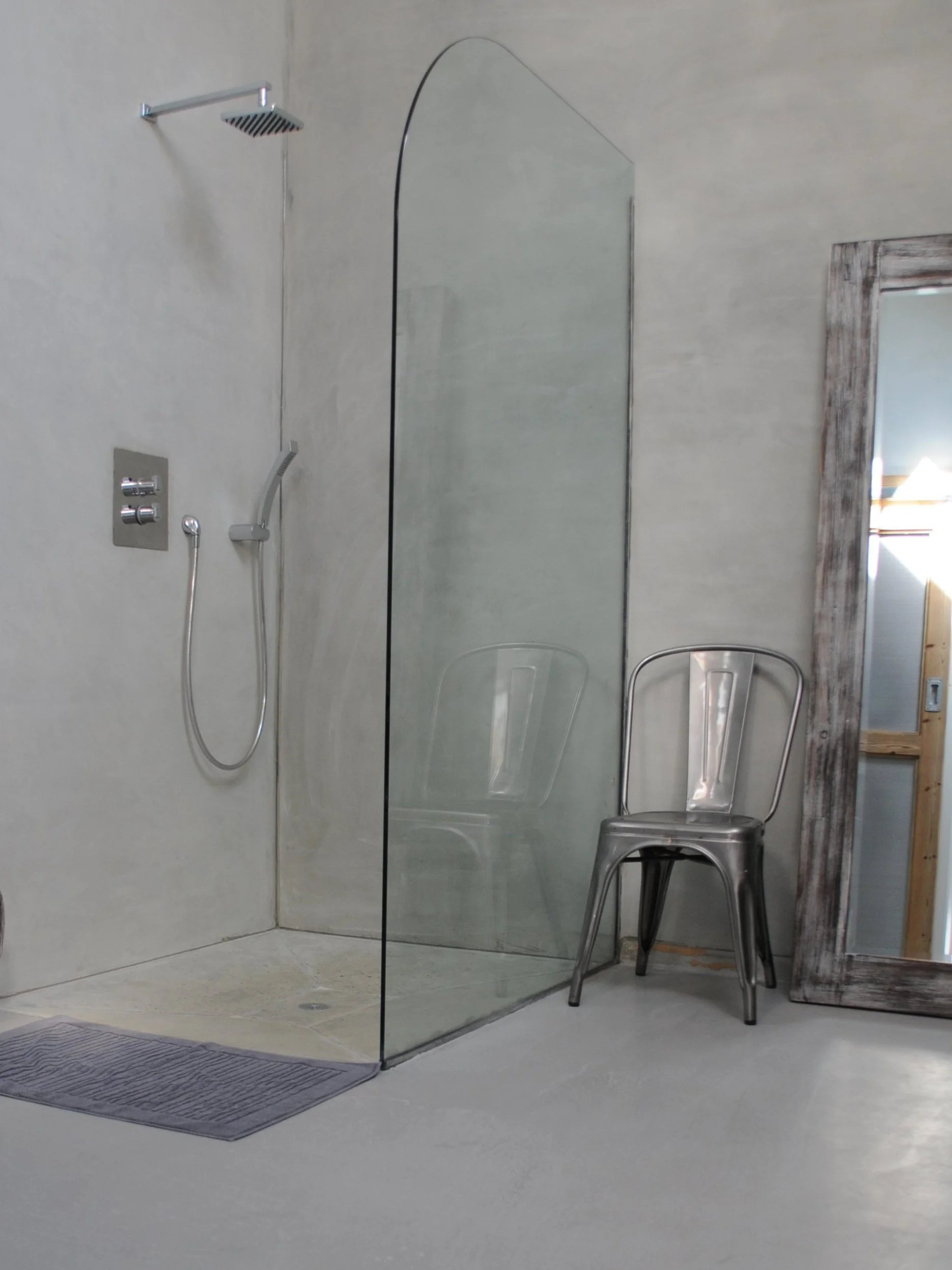 A modern shower area with a glass shower screen, rain showerhead, handheld shower, and a stool, with a rustic wooden-framed mirror nearby.