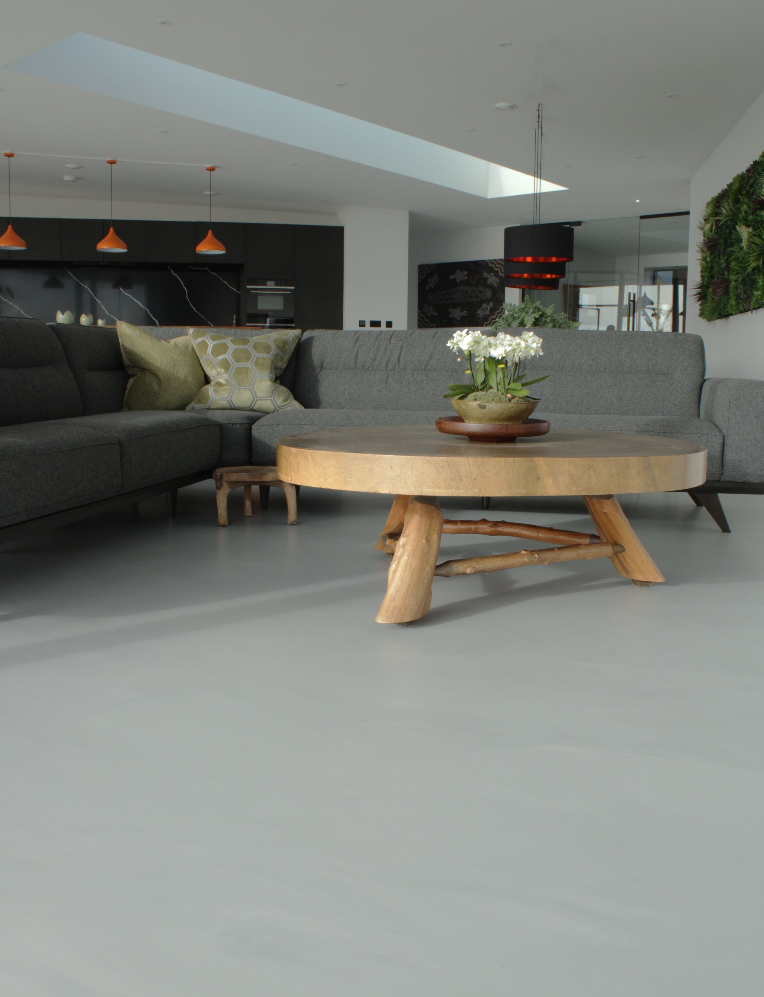 Modern living room with a gray sectional sofa, decorative pillows, wooden coffee table with a flower arrangement, black kitchen cabinets, orange pendant lights, and large windows.