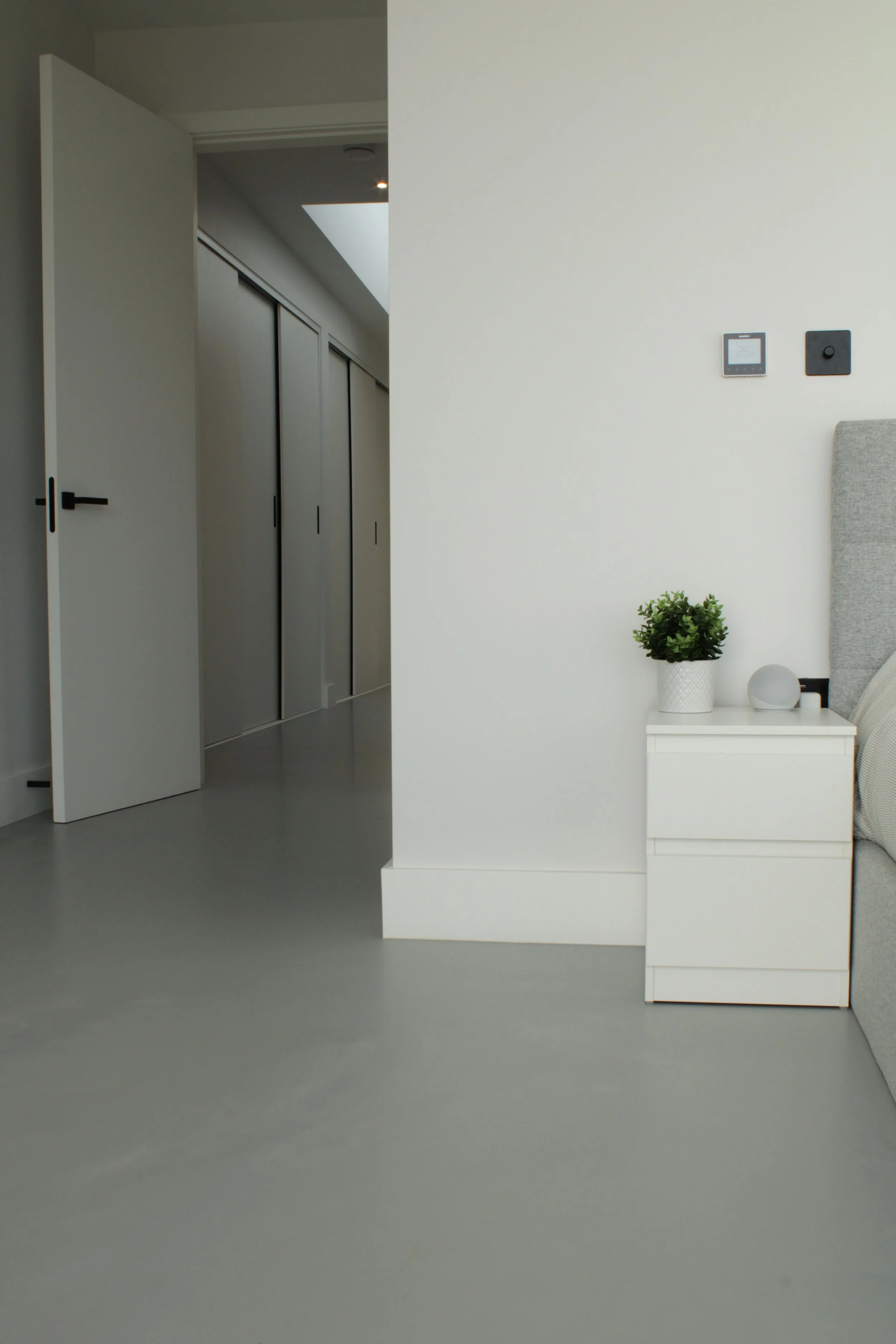 Minimalist interior with white walls, a white nightstand with a green potted plant and a spherical decor item, partially visible bed, digital thermostat, black switch on wall, gray flooring, open door leading to hallway with closet doors.