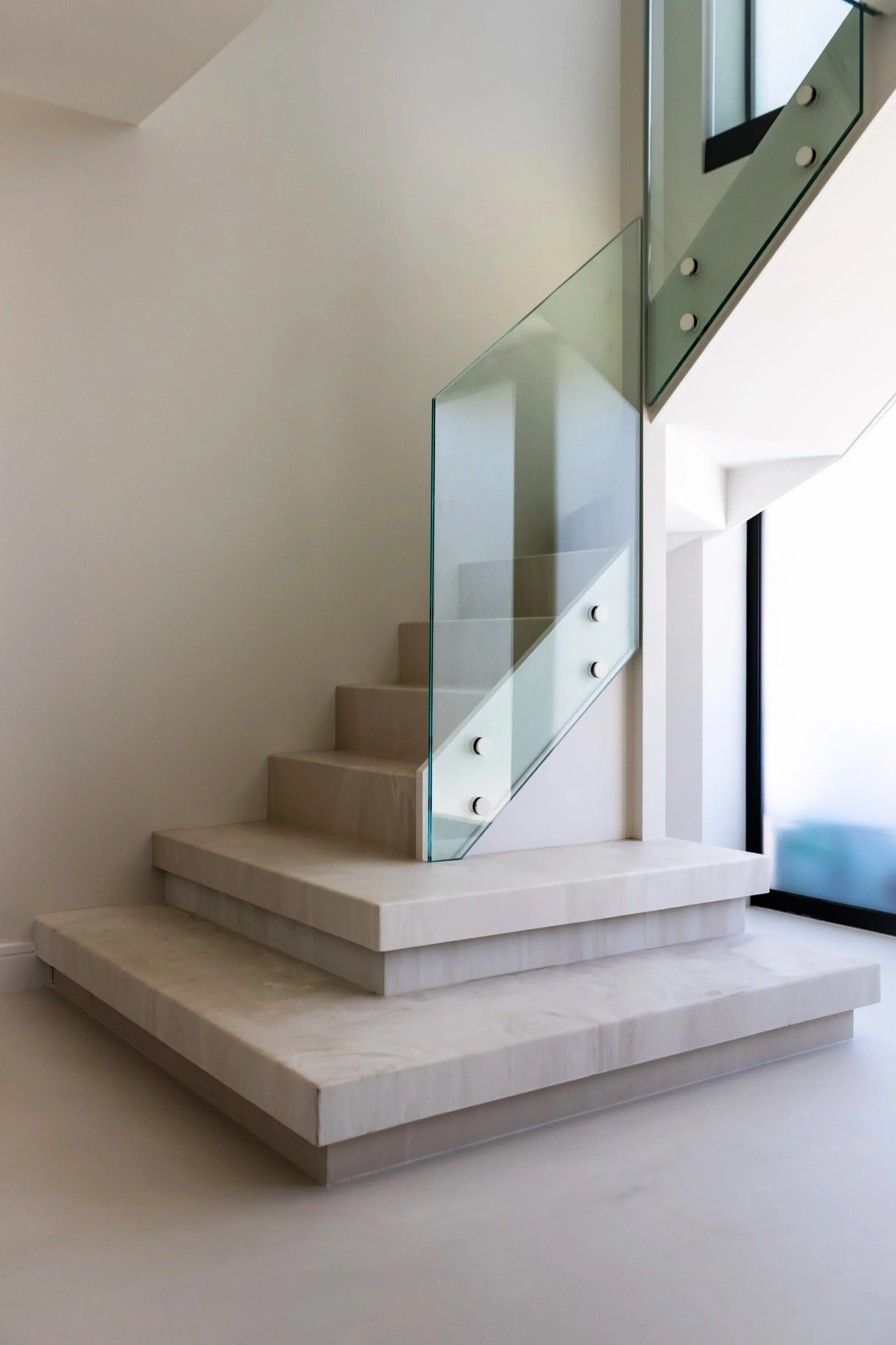 Modern staircase with beige steps and a glass railing inside a minimalist building, near large windows.