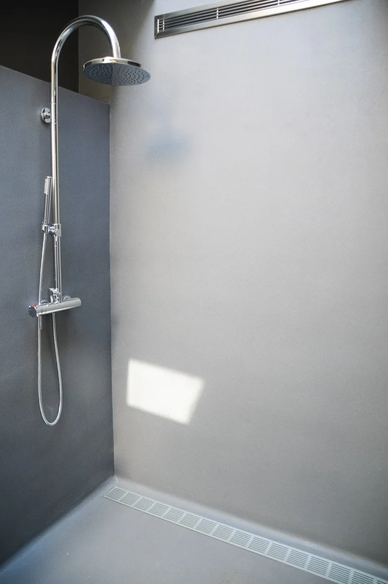Modern shower with a chrome showerhead, sliding control, and a drain on the floor. Light-colored walls with a vent on the upper wall.