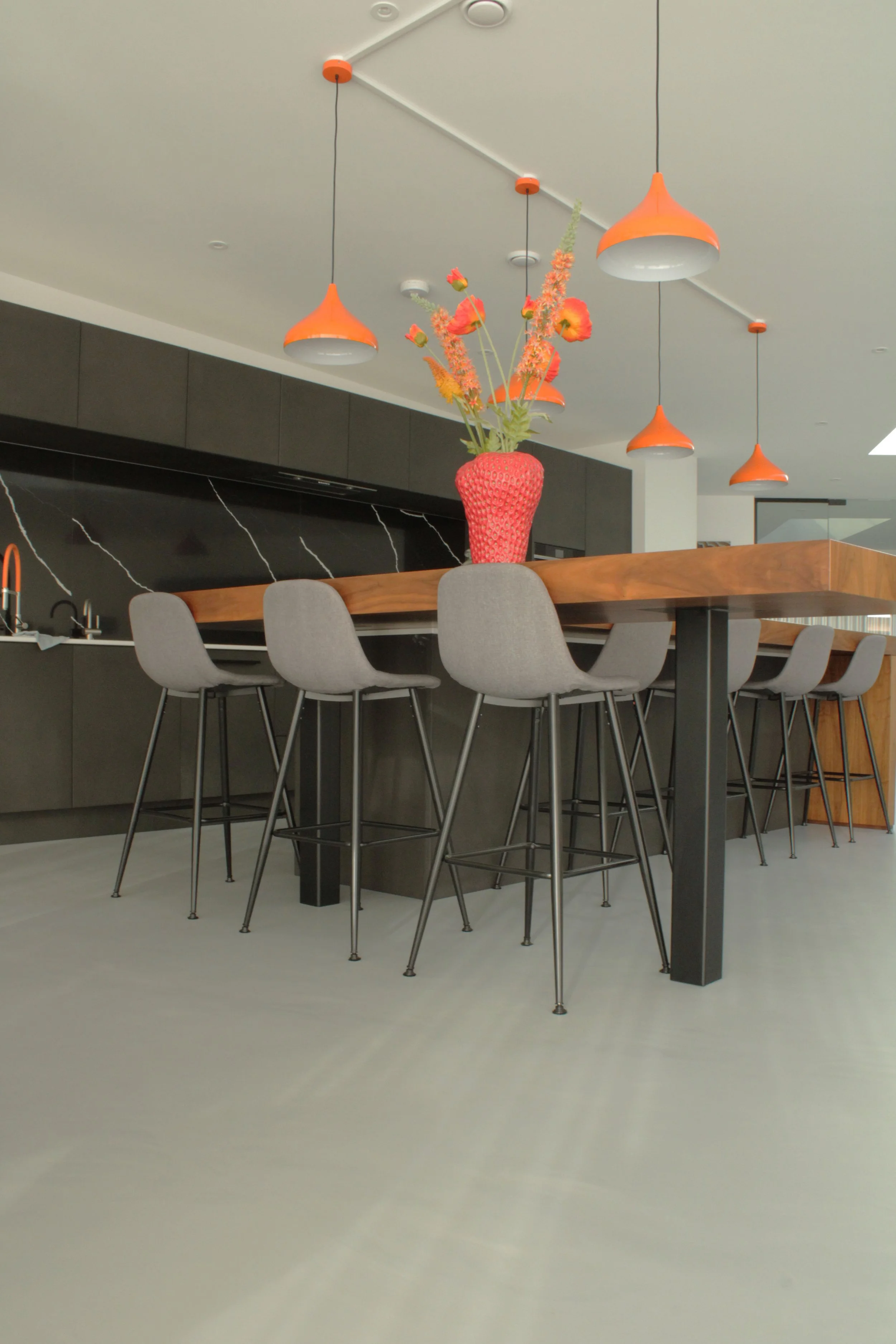 A modern kitchen with a black marble backsplash, wooden table, gray chairs, and hanging orange pendant lights. A red vase with orange and pink flowers is placed on the table.
