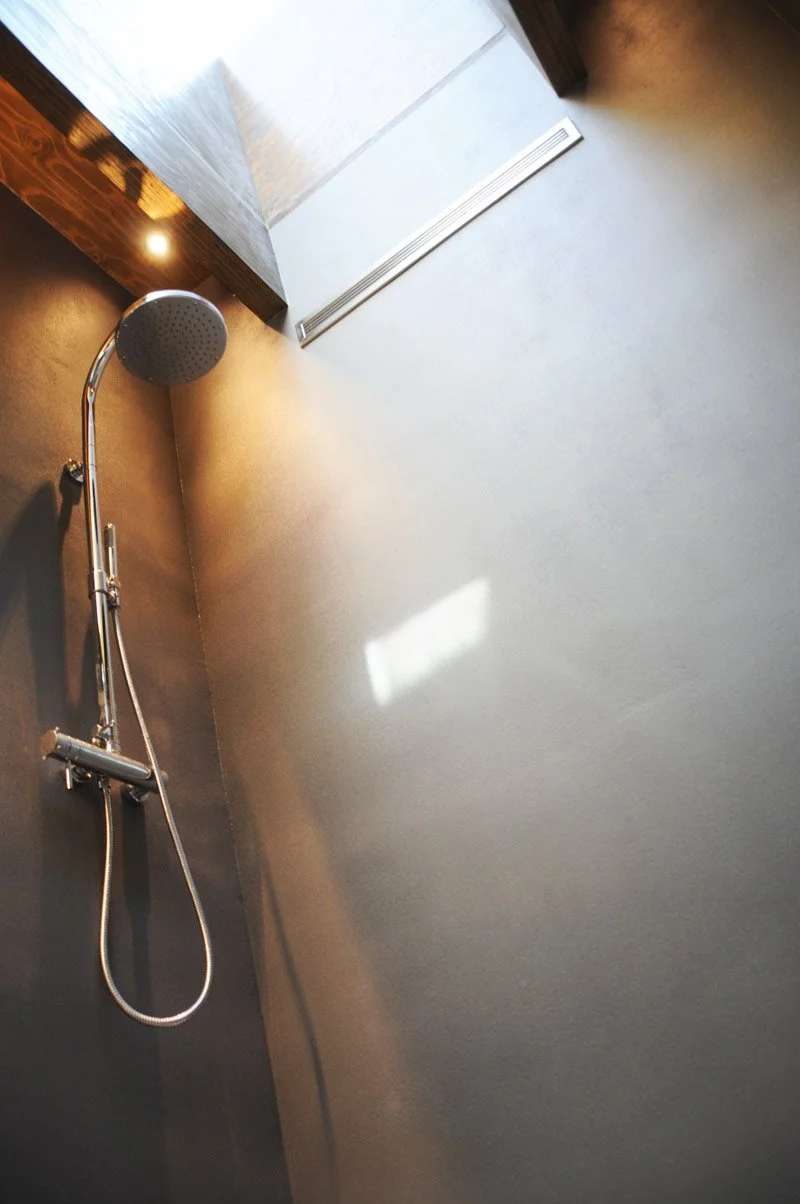 A modern shower with a mounted showerhead and adjustable hose, situated next to a dark brown wall and a receptacle vent, with natural light coming from a window.