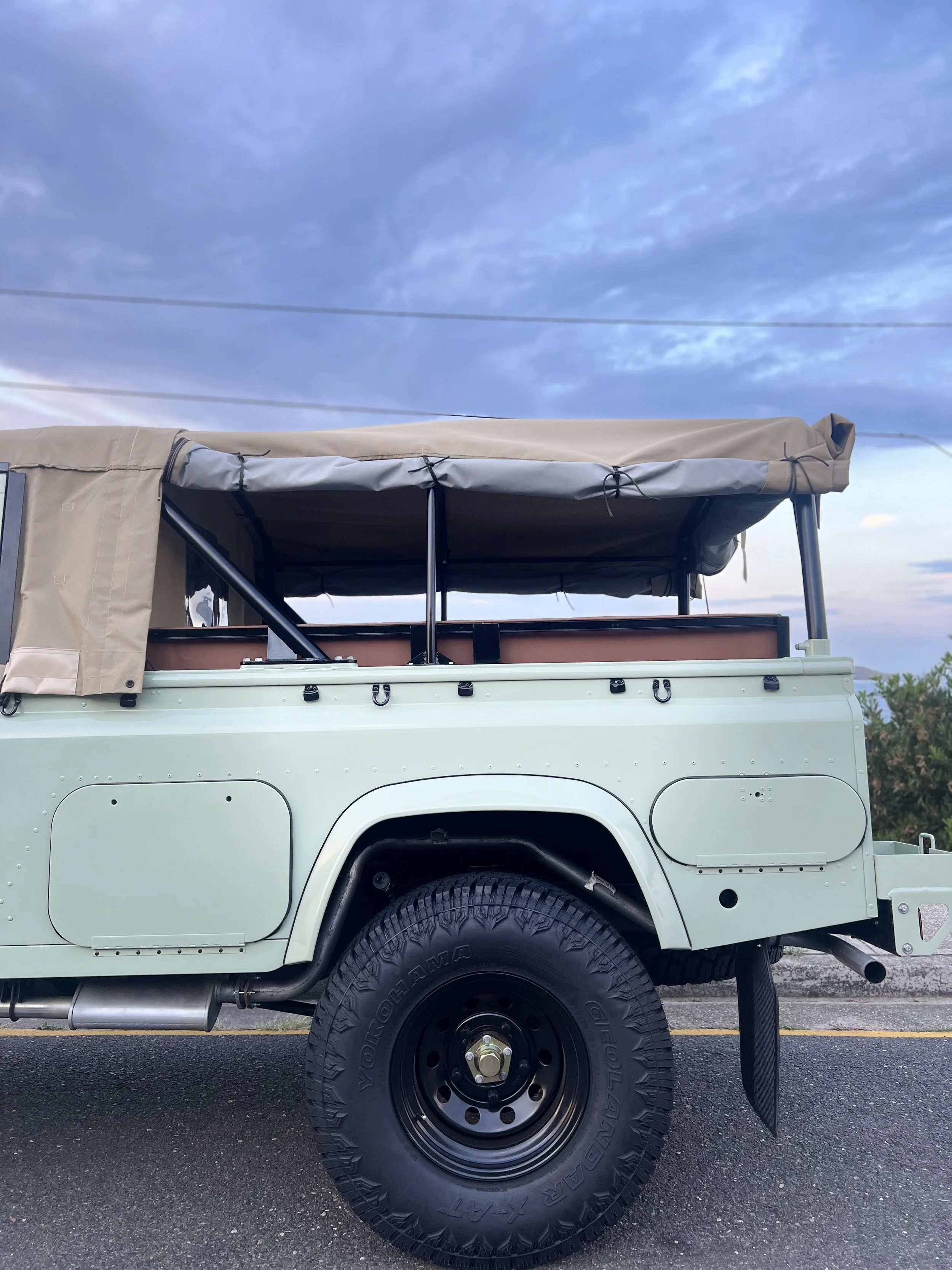 A close-up side view of a beige off-road vehicle with a roof rack, large all-terrain tire, and partial open storage compartments, parked on an asphalt surface with a cloudy sky in the background.