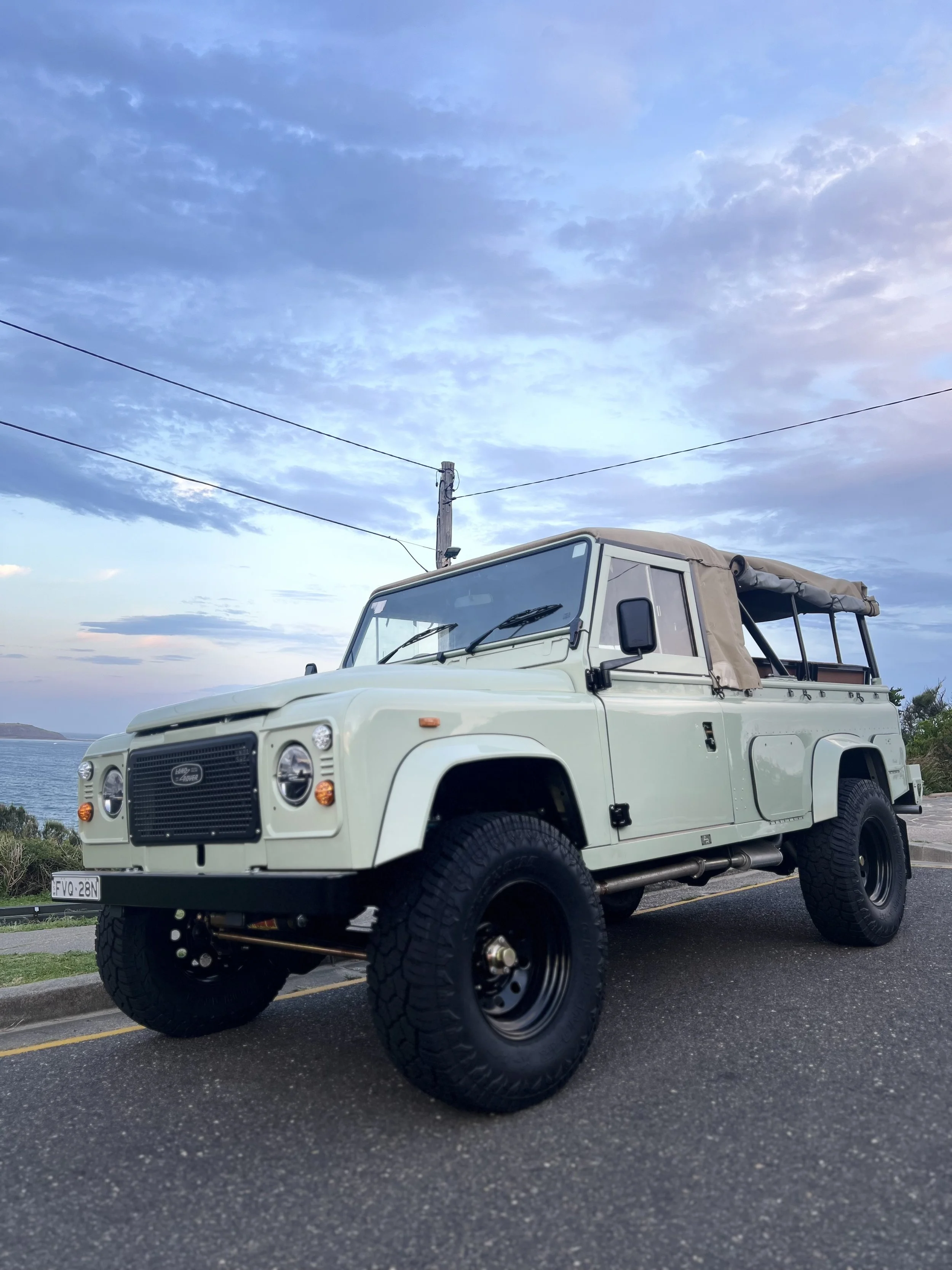 A white, vintage off-road vehicle with large black tires parked on the street near a coastal area, with a utility pole and wires overhead and water visible in the background under a partly cloudy sky.