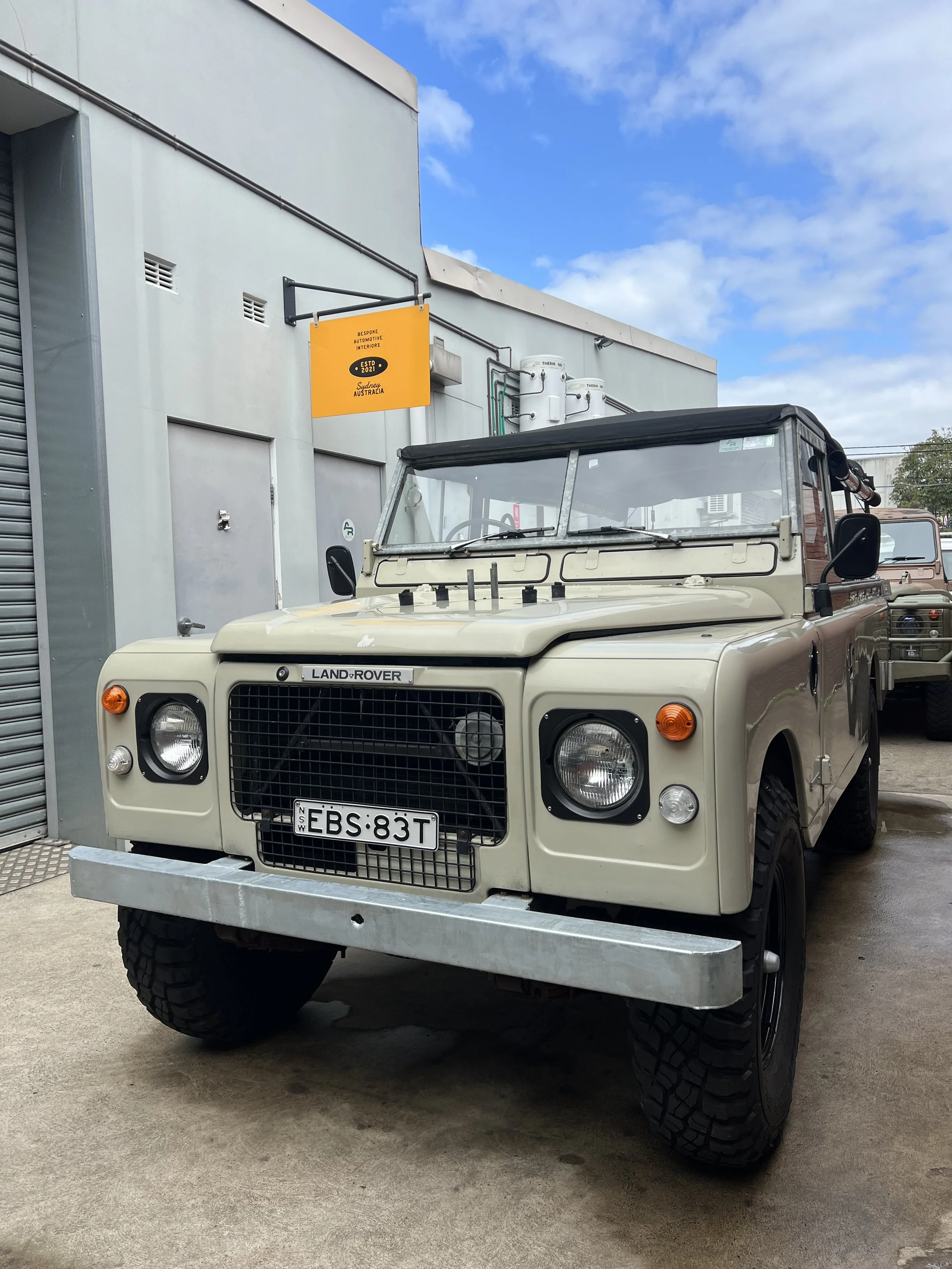 A vintage beige Land Rover parked outside a building with a white wall and a yellow sign that reads 'Bespoke Automotive Interiors, Estd 2021, Sydney, Australia.' The vehicle has large off-road tires, black side mirrors, and a license plate that reads