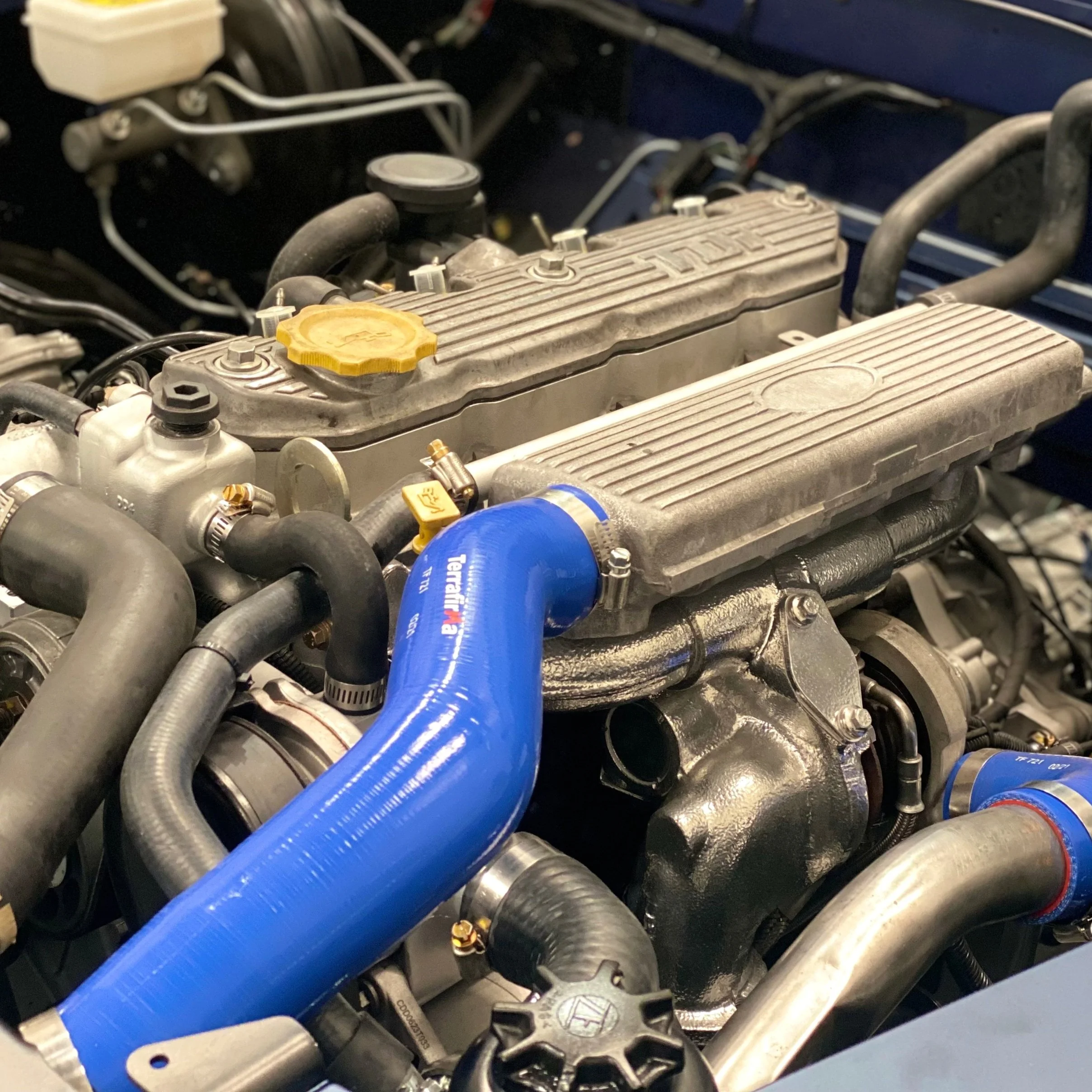 Close-up photo of a car engine showing various hoses, clamps, and metallic components, including a prominent blue silicone hose.