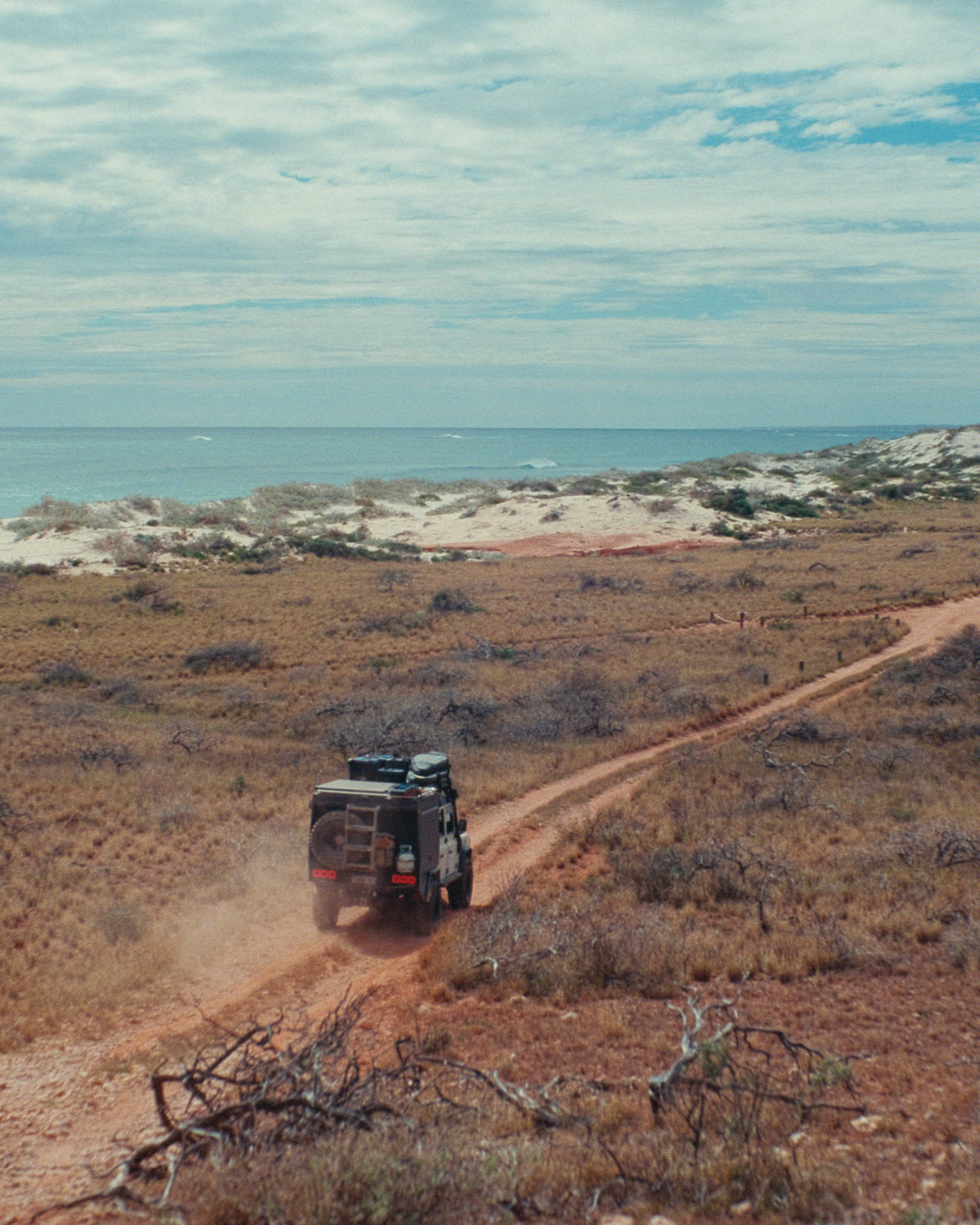 A Jeep driving on a dirt trail through a dry, grassy landscape with sparse bushes. In the background, there is an ocean or sea and a partly cloudy sky.