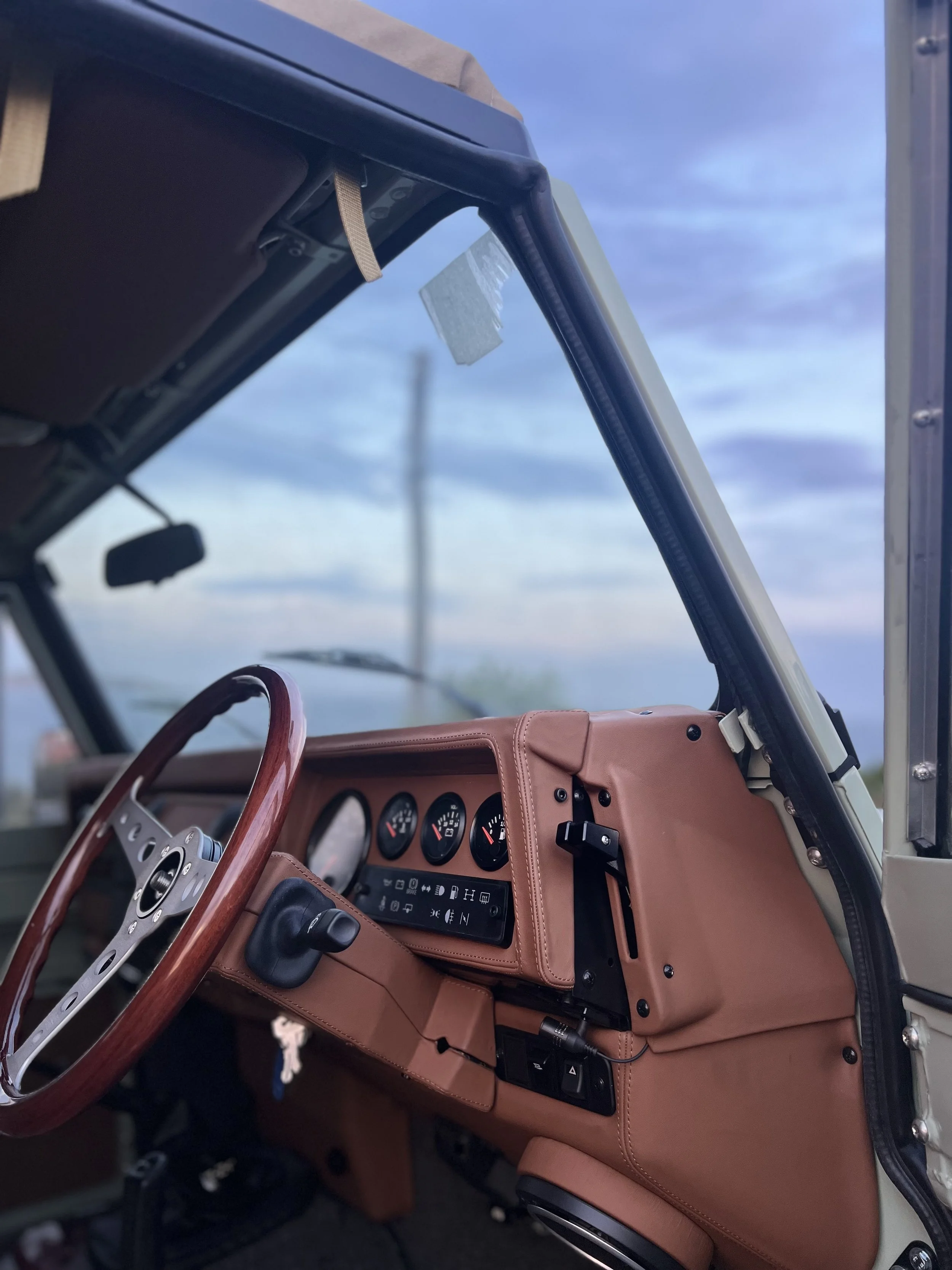 Close-up of the interior of a vintage vehicle, showcasing a wooden steering wheel, a tan dashboard with gauges, switches, and a black cable, with a clear blue sky in the background.