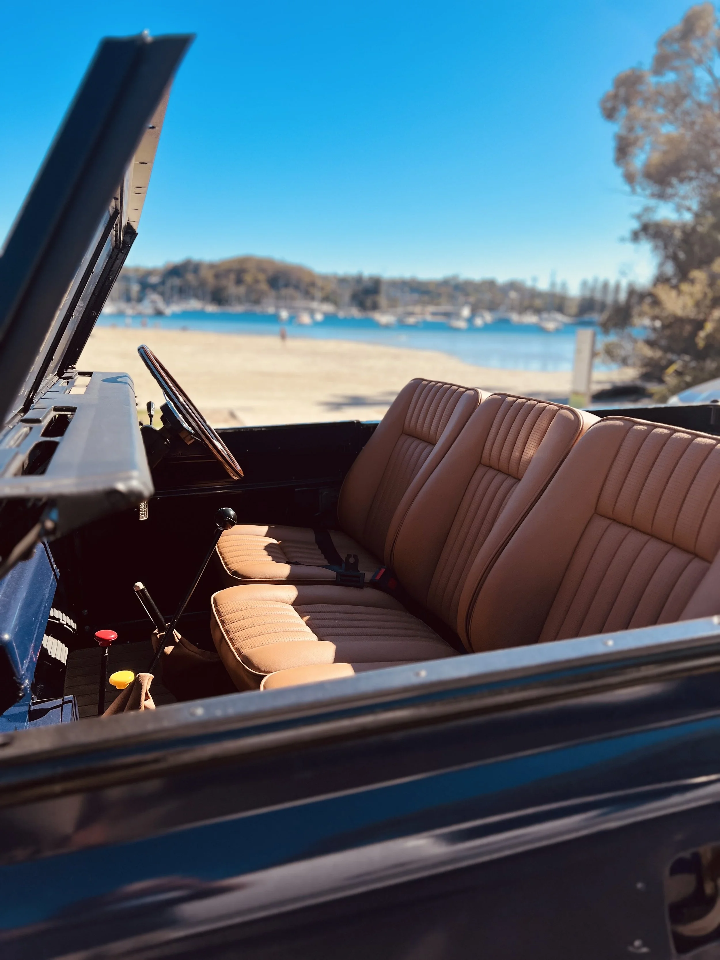 View from inside a vintage vehicle showing tan leather seats and an open hood, with a scenic outdoor background including a beach, water, boats, trees, and a clear blue sky.