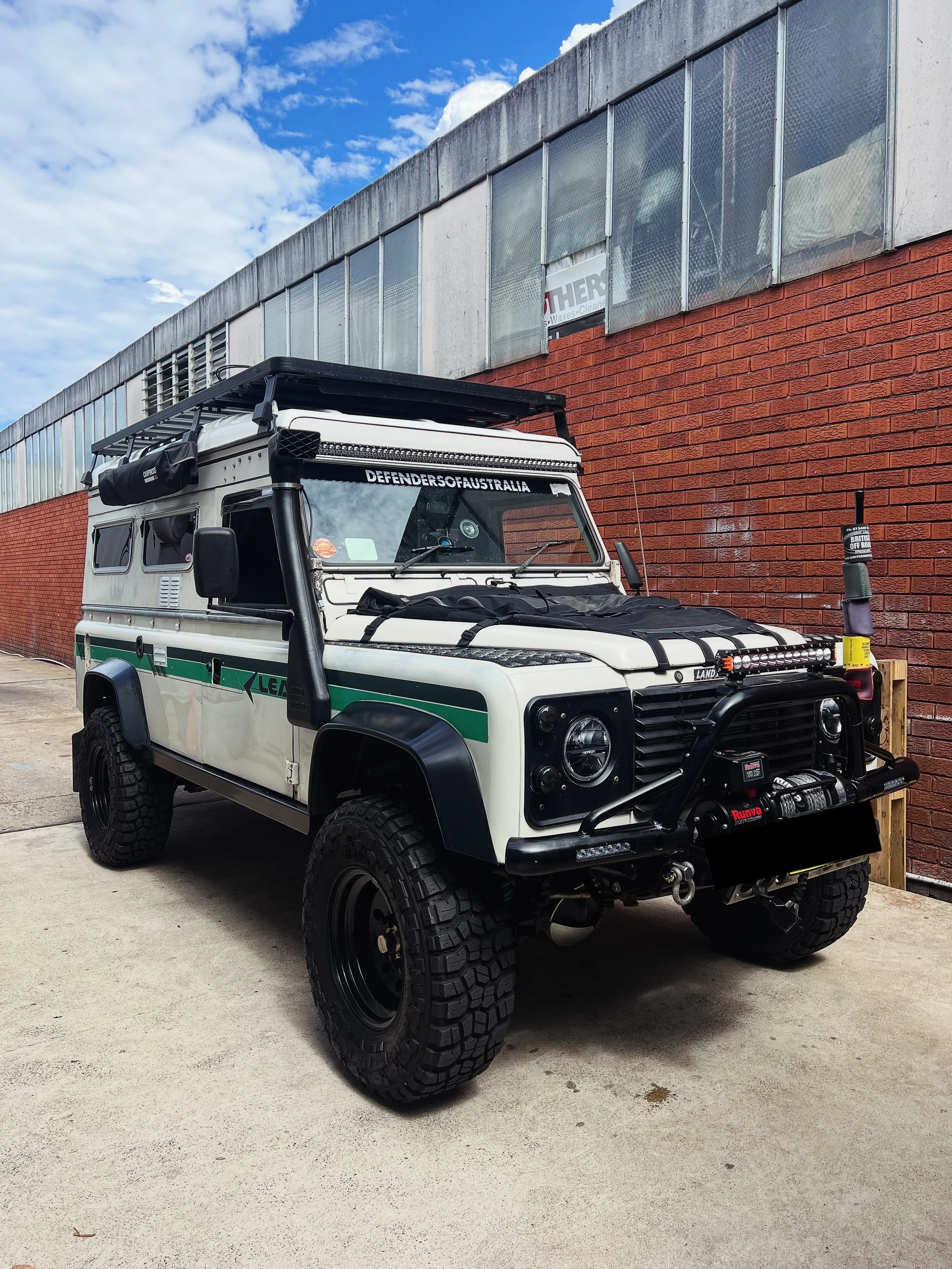 A white off-road vehicle with black tires and accessories parked outdoors next to a brick wall and a modern building with metallic panels. The vehicle features a roof rack, front bumper, and a sticker that says 'DEFENDERSOFAUSTRALIA'. The sky is part