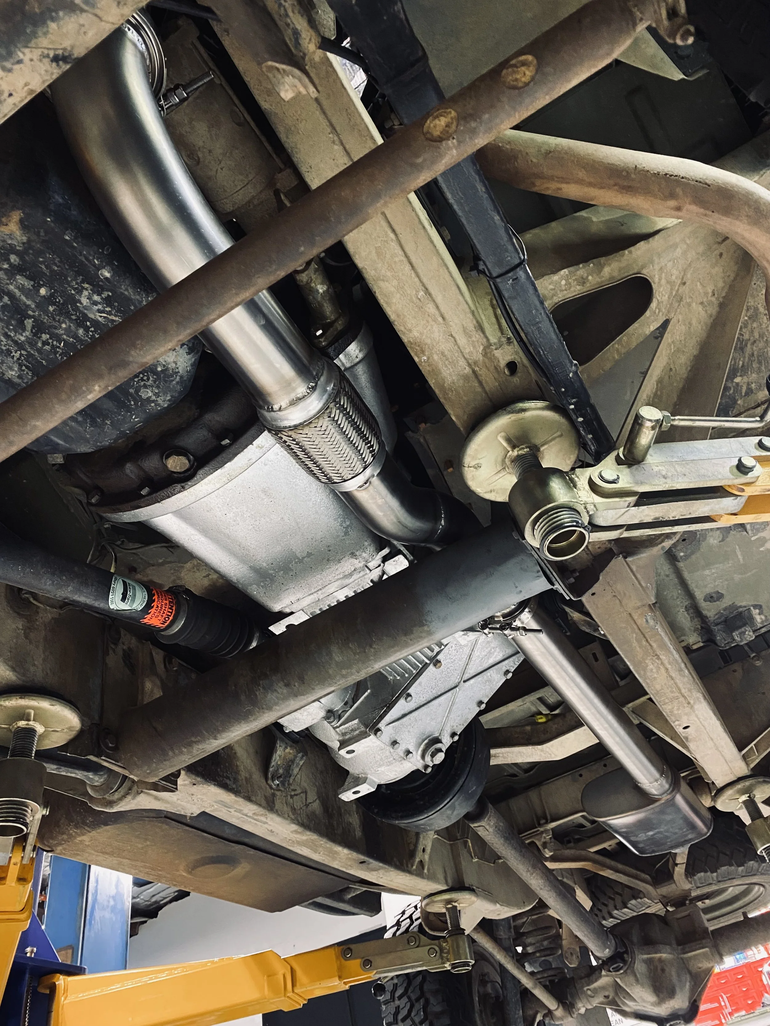Underneath a vehicle showing the exhaust system, driveshaft, and suspension parts in a garage or workshop.