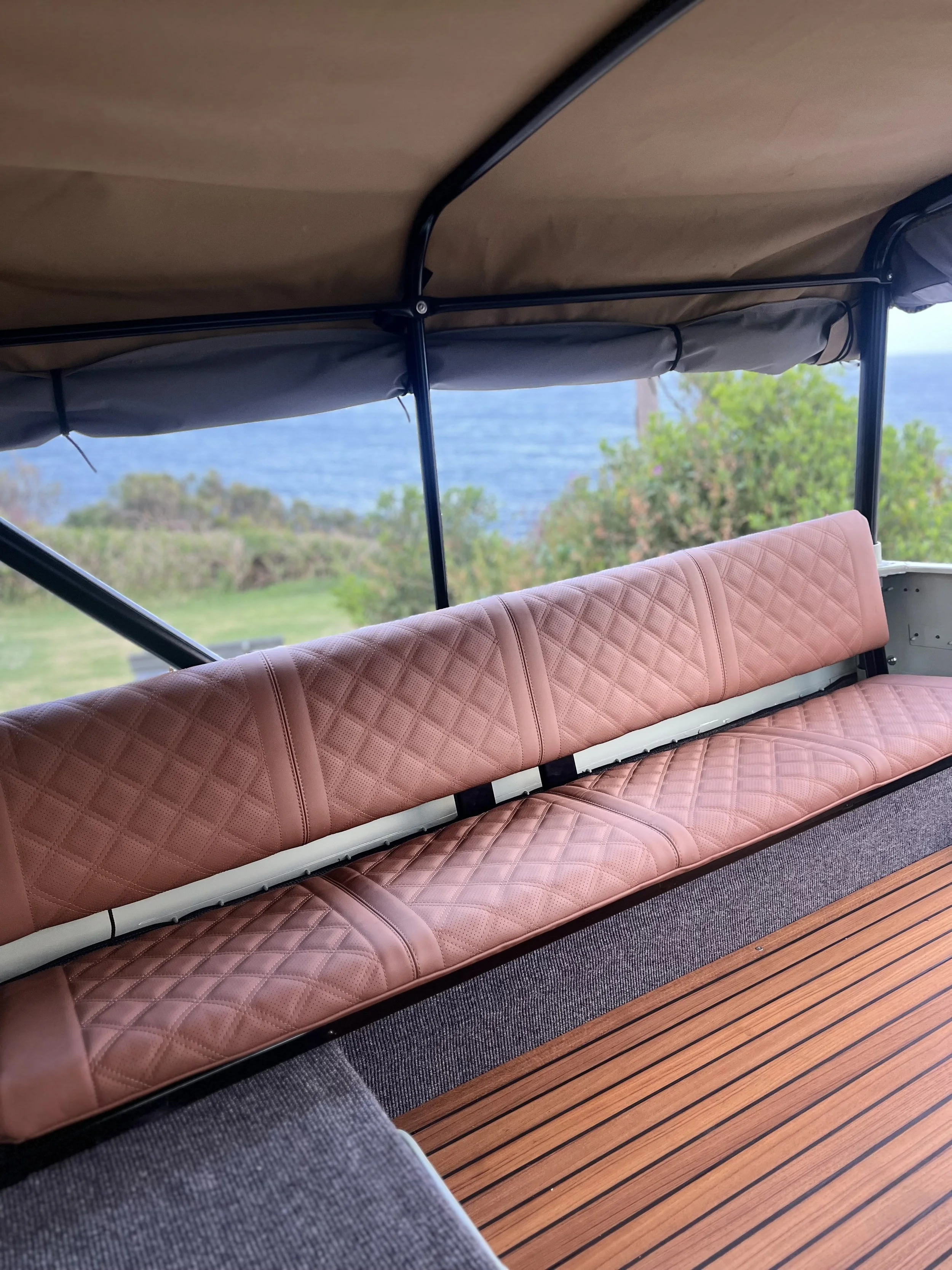 Inside a boat with pink quilted leather bench seats, a wooden floor, and an open view of a grassy landscape with trees and water in the distance.