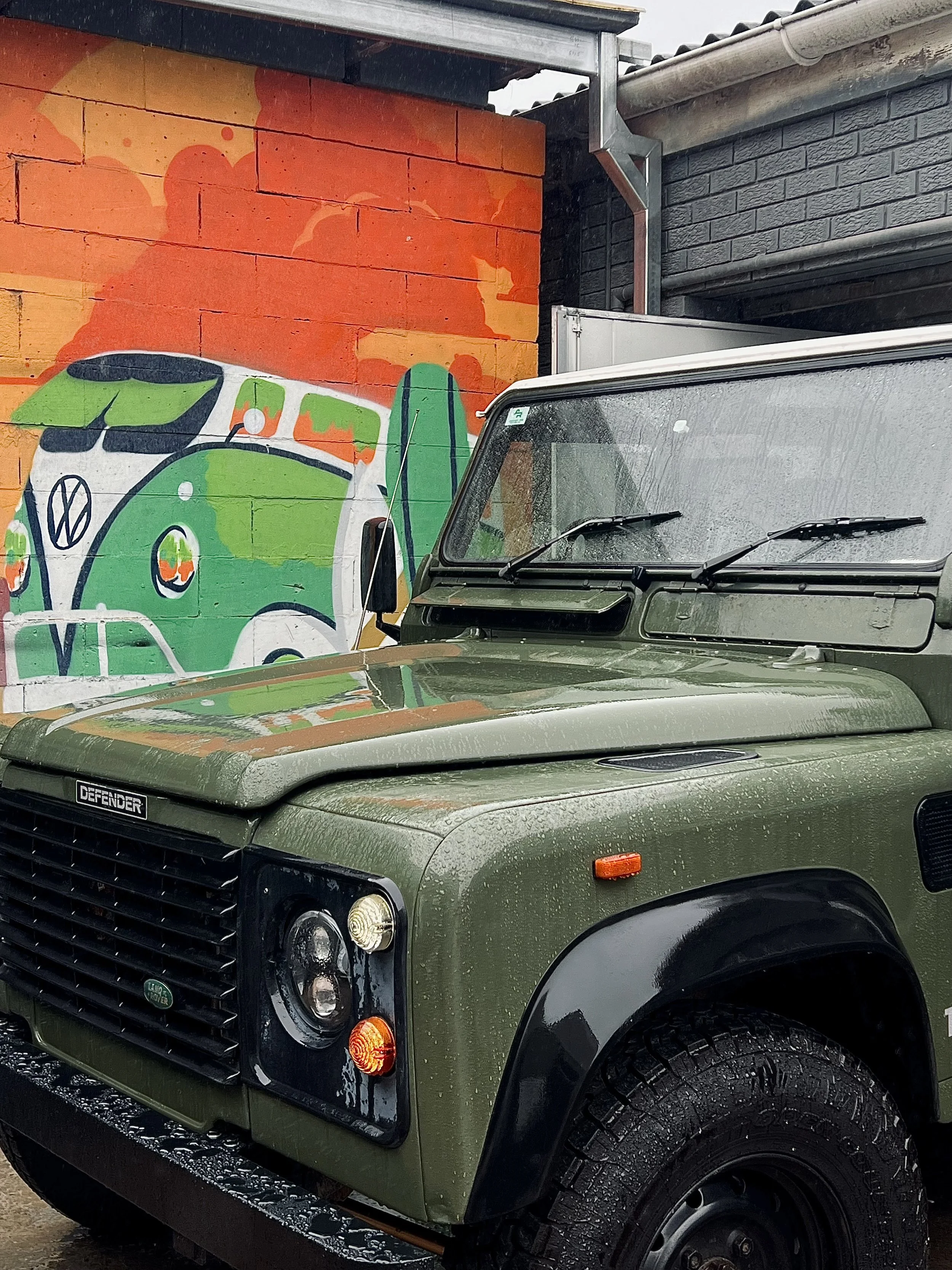 Close-up view of a matte green Land Rover Defender vehicle with rain droplets on it, parked outdoors next to a brick wall art mural featuring a colorful illustration of a green and white camper van on an orange and red background.