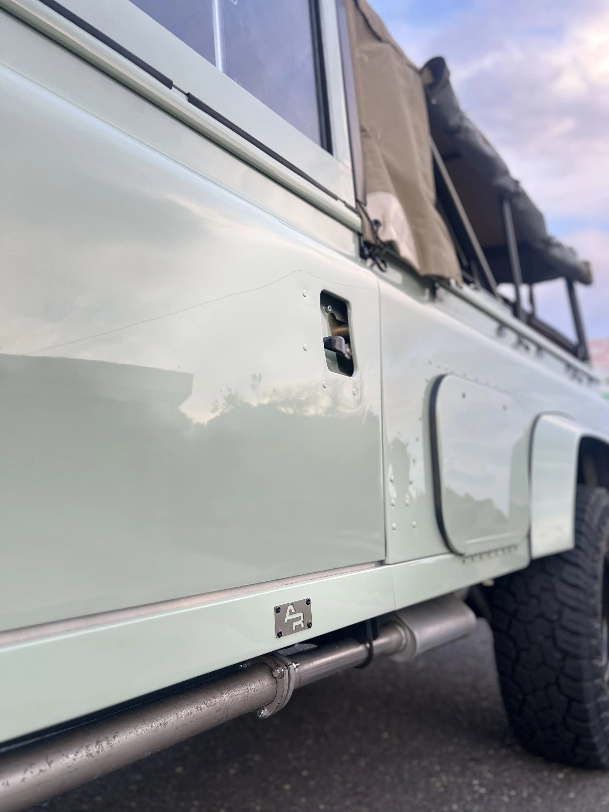 Close-up of the side of a light-colored off-road vehicle, showing the door handle, a step with a label, and part of a tire with a rugged tread pattern. The vehicle has a roof rack with items stored on top and a side robot guard.