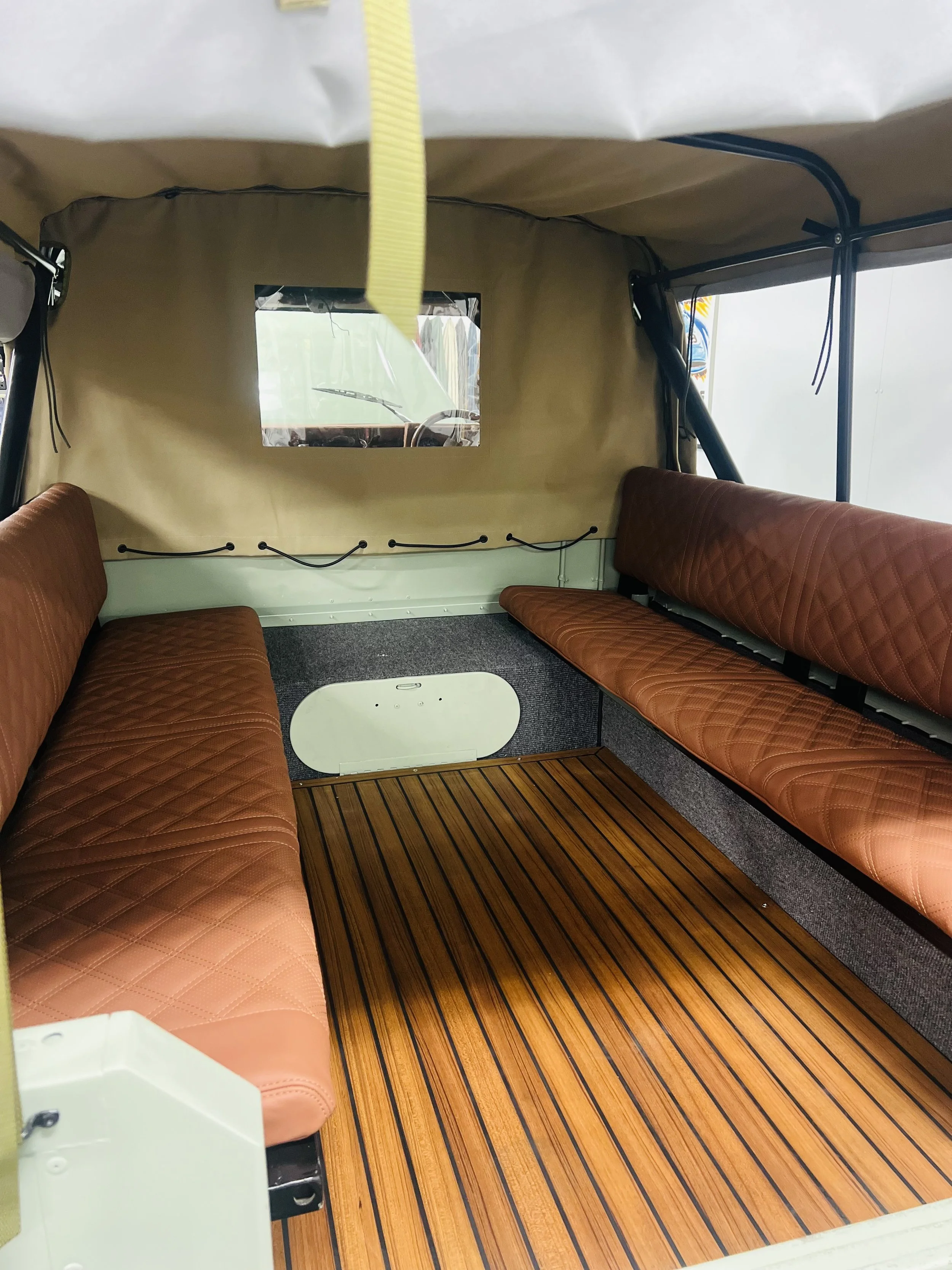 Interior of a vintage vehicle with two brown leather bench seats, a striped wooden floor, a small oval table, and a beige fabric wall with a rectangular window showing the vehicle's dashboard and steering wheel.