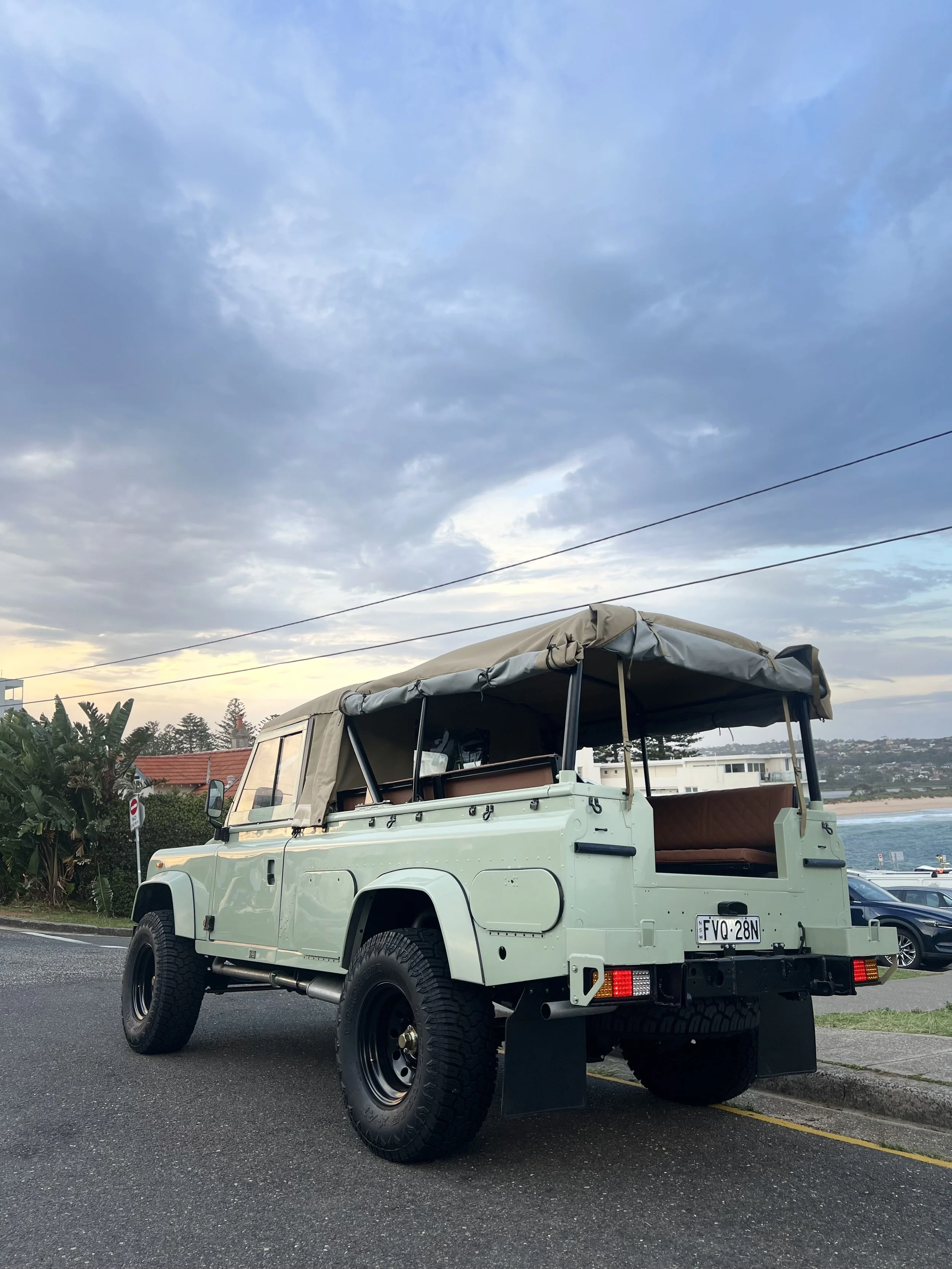 Light green vintage off-road vehicle with a beige canvas roof parked on the street with ocean and sky in the background.