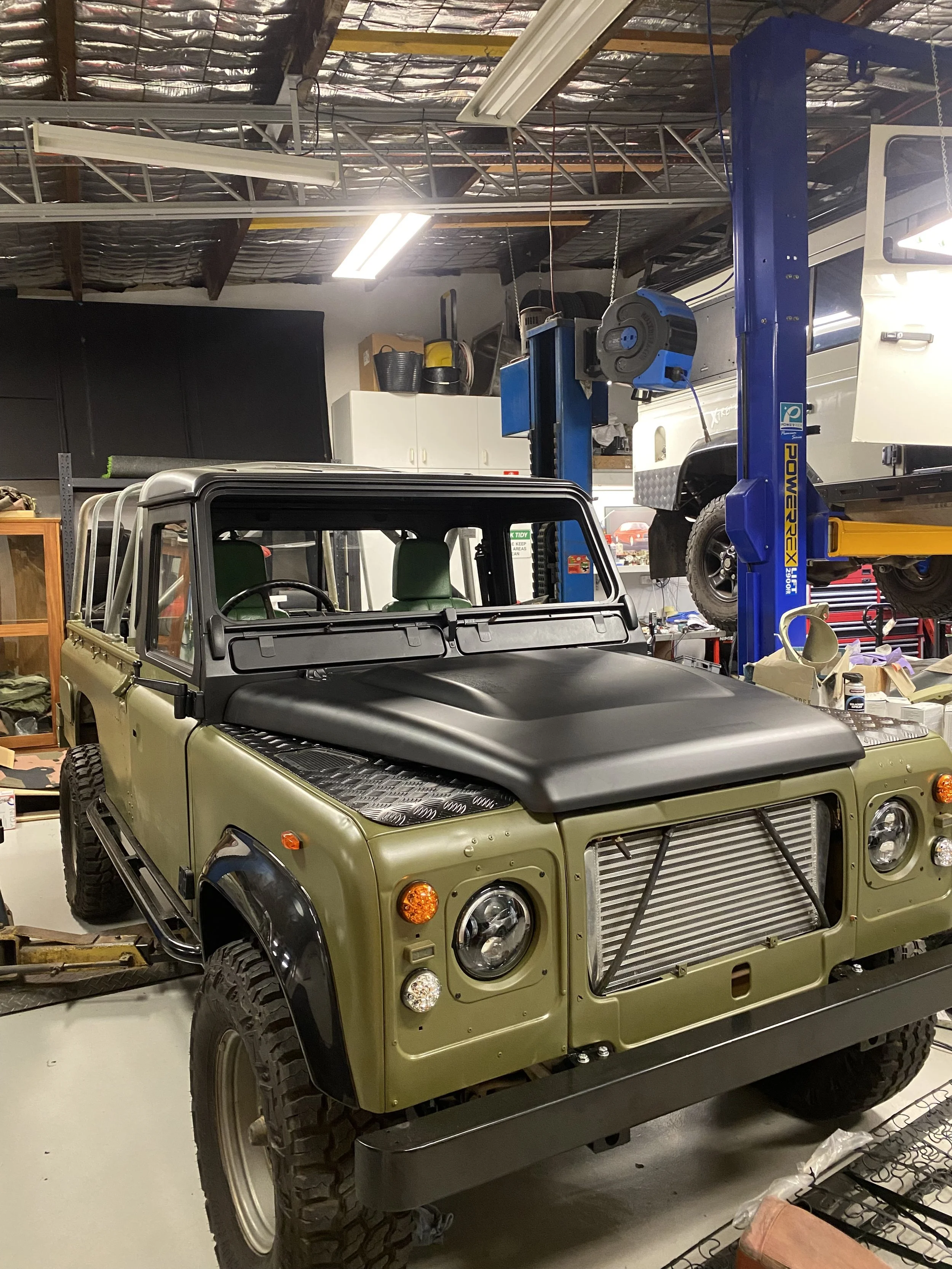 An unfinished off-road vehicle, resembling a Land Rover Defender, is in a workshop with a lift holding a vehicle above. The workshop has various tools, cabinets, and equipment.