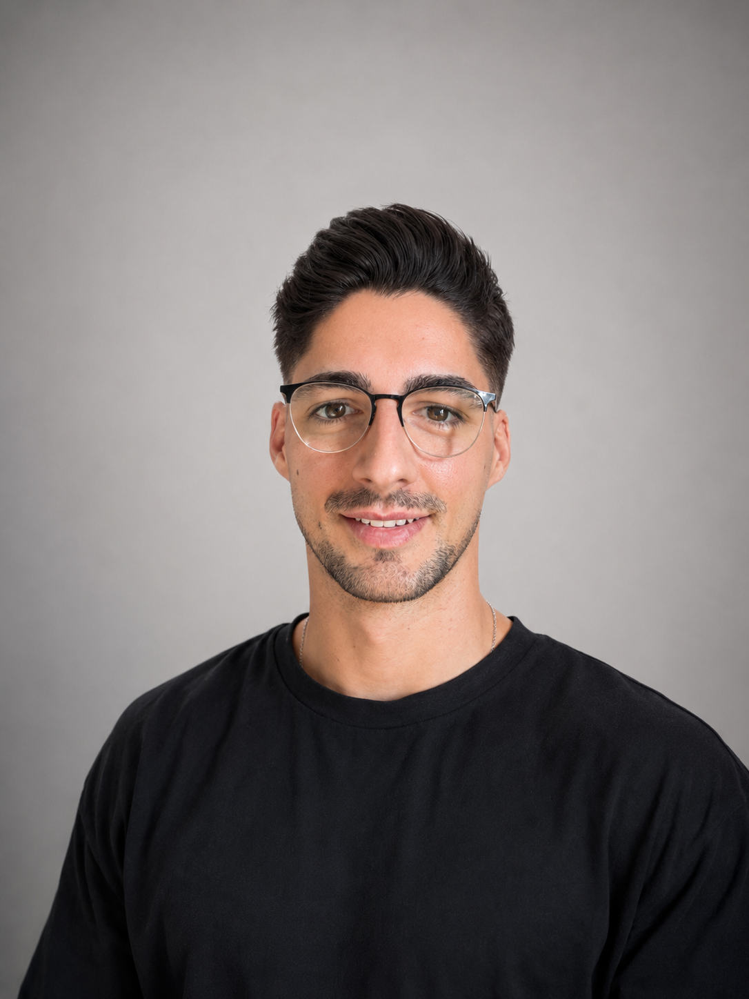 Porträt eines jungen Mannes mit dunklen Haaren, Brille und schwarzem T-Shirt vor grauem Hintergrund.