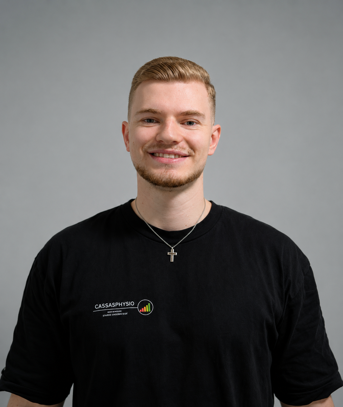 Junger Mann mit blondem Haar, Bart und Kreuzkette, trägt schwarzes T-Shirt mit Logo und Text, lächelt vor grauem Hintergrund.
