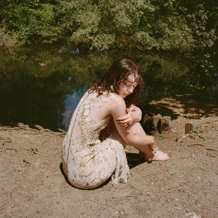A woman sitting on the ground near a body of water, surrounded by trees, with curly dark hair and wearing a light, patterned dress.
