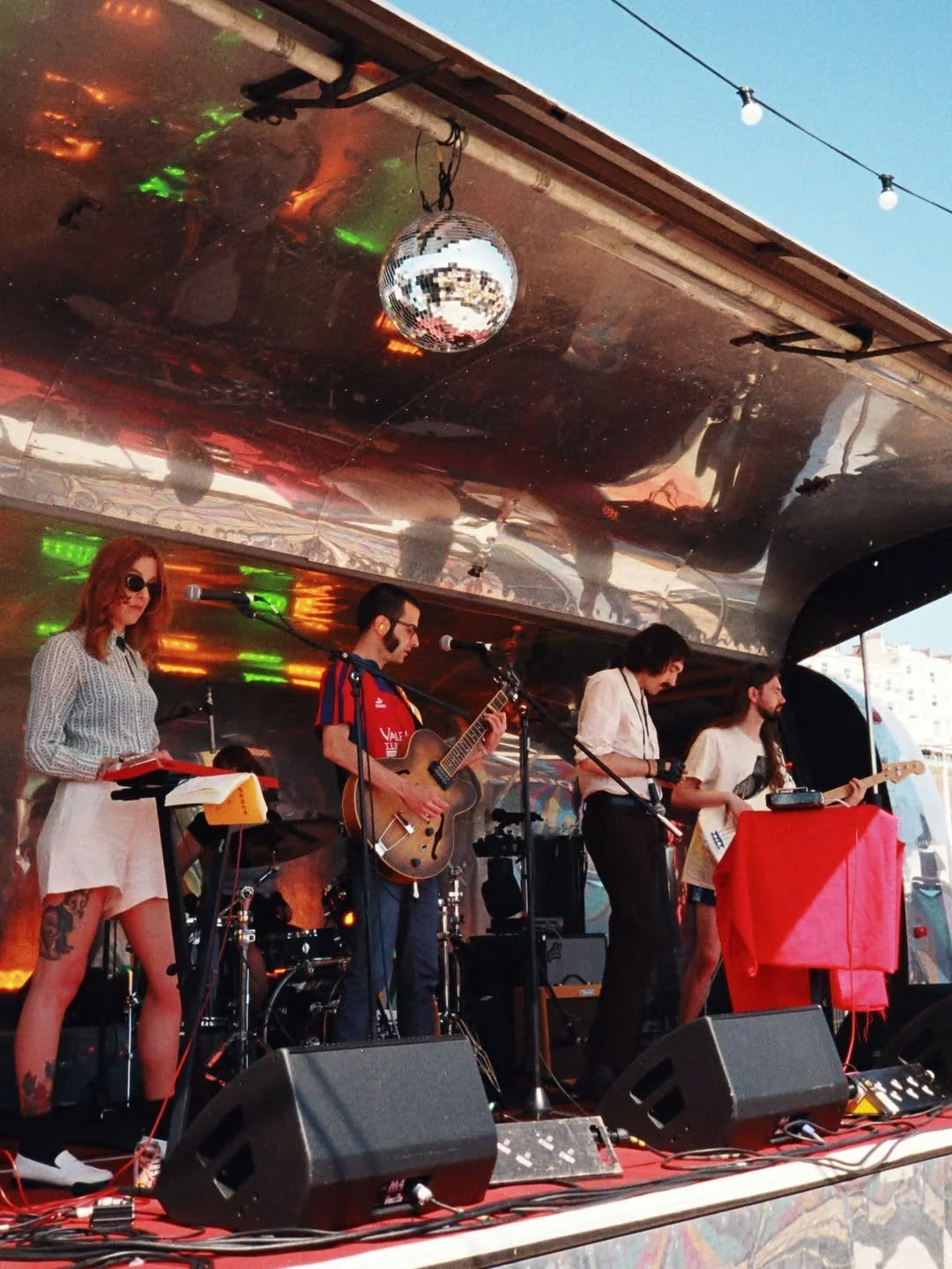 A band performing live on stage at an outdoor event with a disco ball hanging from the ceiling, a woman with red hair and sunglasses, and four band members playing instruments.