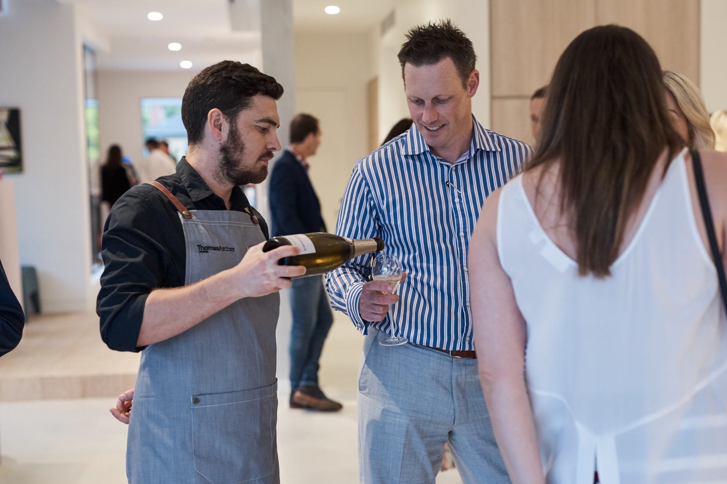 A man in a gray apron is pouring a drink into a glass held by another man in a striped shirt at a social event, with other people in the background.