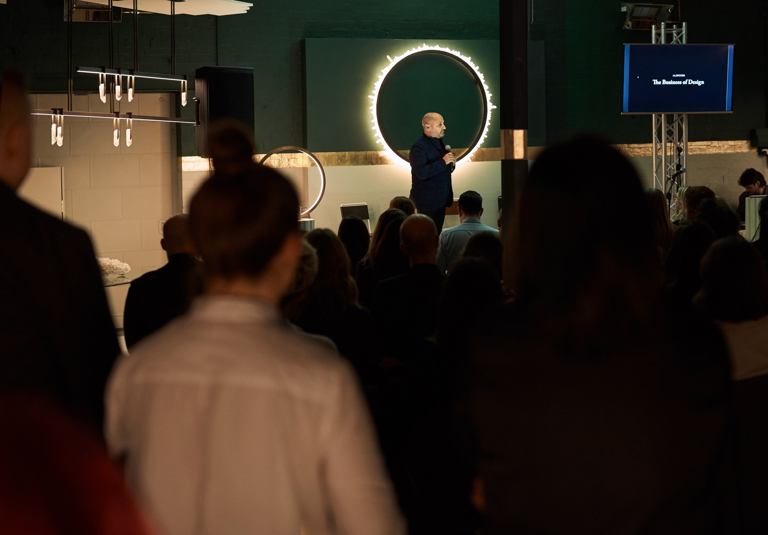 Speaker giving a presentation in a dimly lit room with audience seated, large ring light behind speaker, screen displaying 'The Business of Design.'