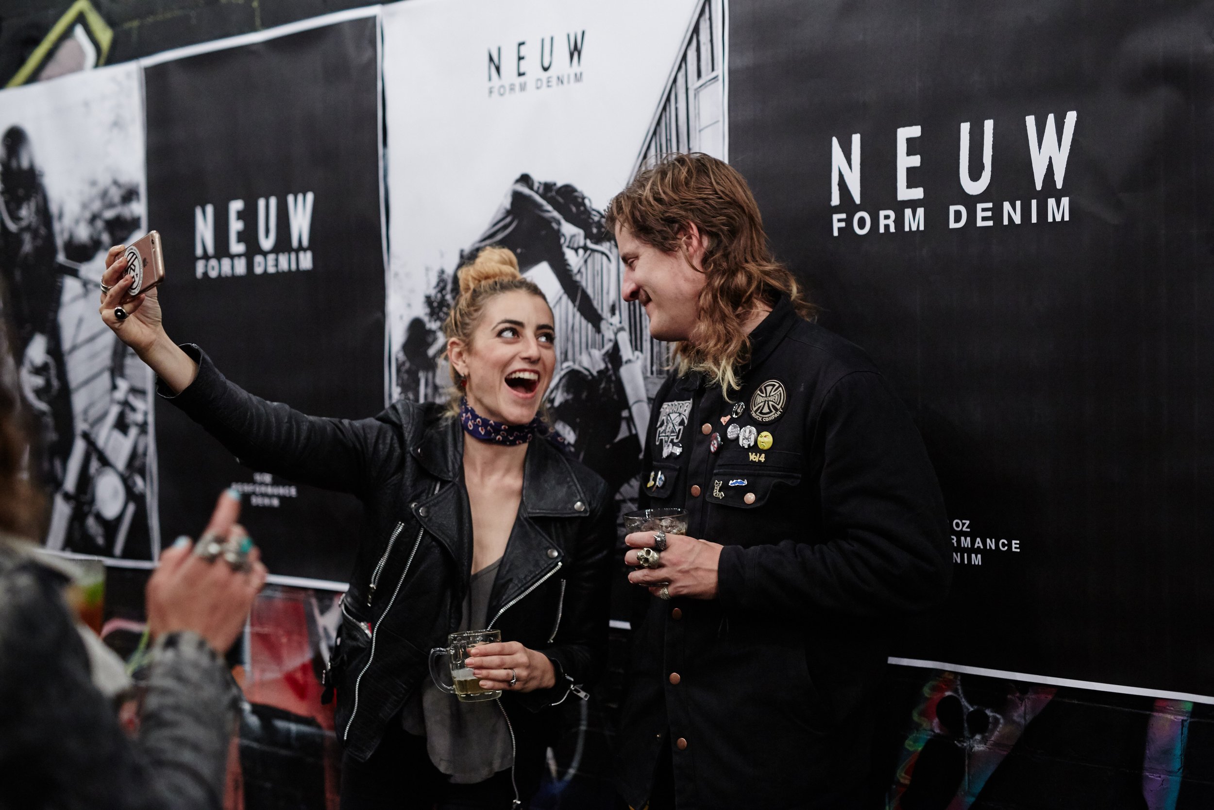Two people taking a selfie at a Neuw Form Denim event, standing in front of promotional posters, both smiling and holding drinks.