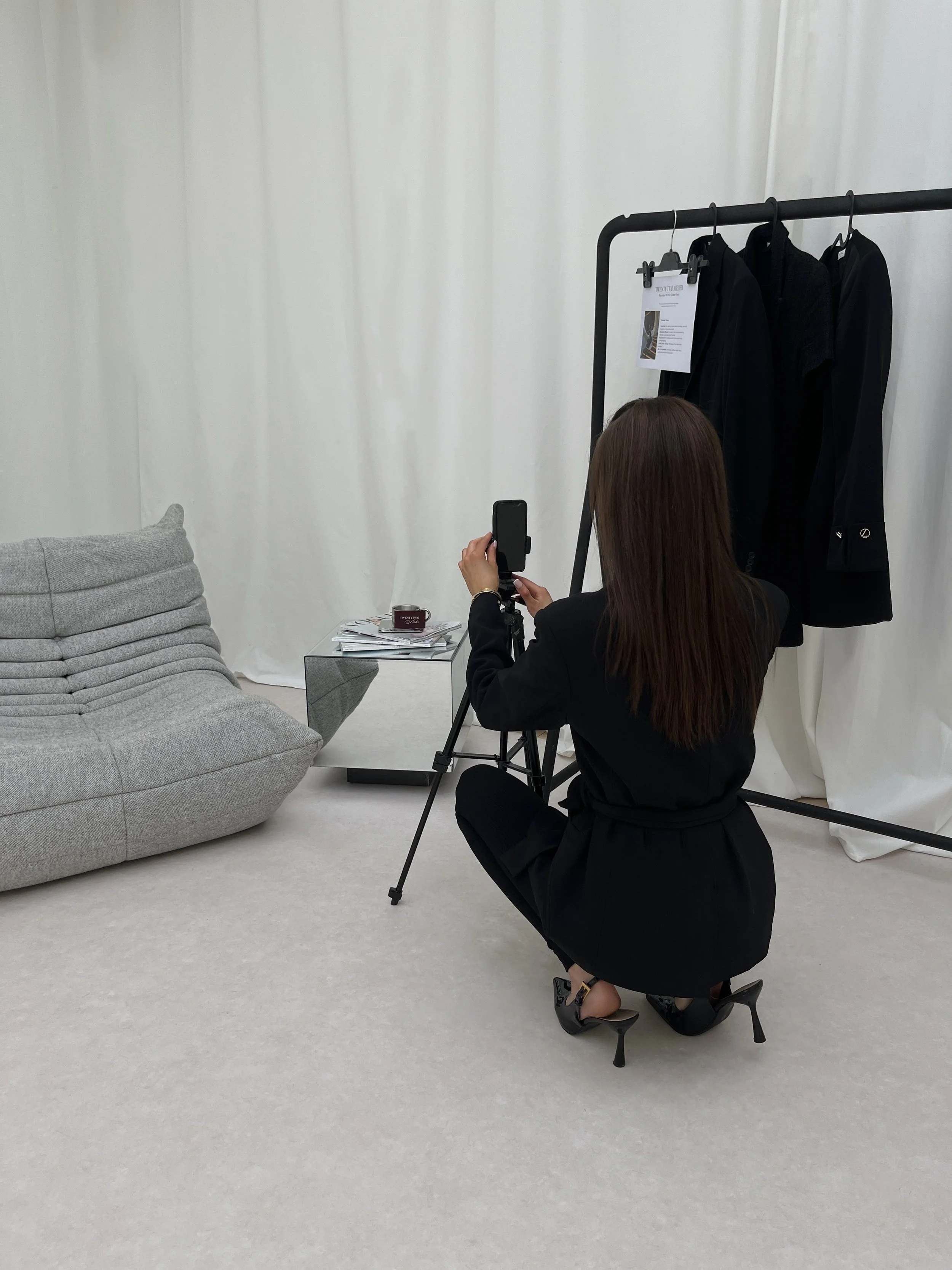 A woman in a black suit squatting on a beige carpet, taking a photo with a smartphone on a tripod in front of black clothing hanging on a black clothing rack, in a minimalist room with white walls and a gray couch.