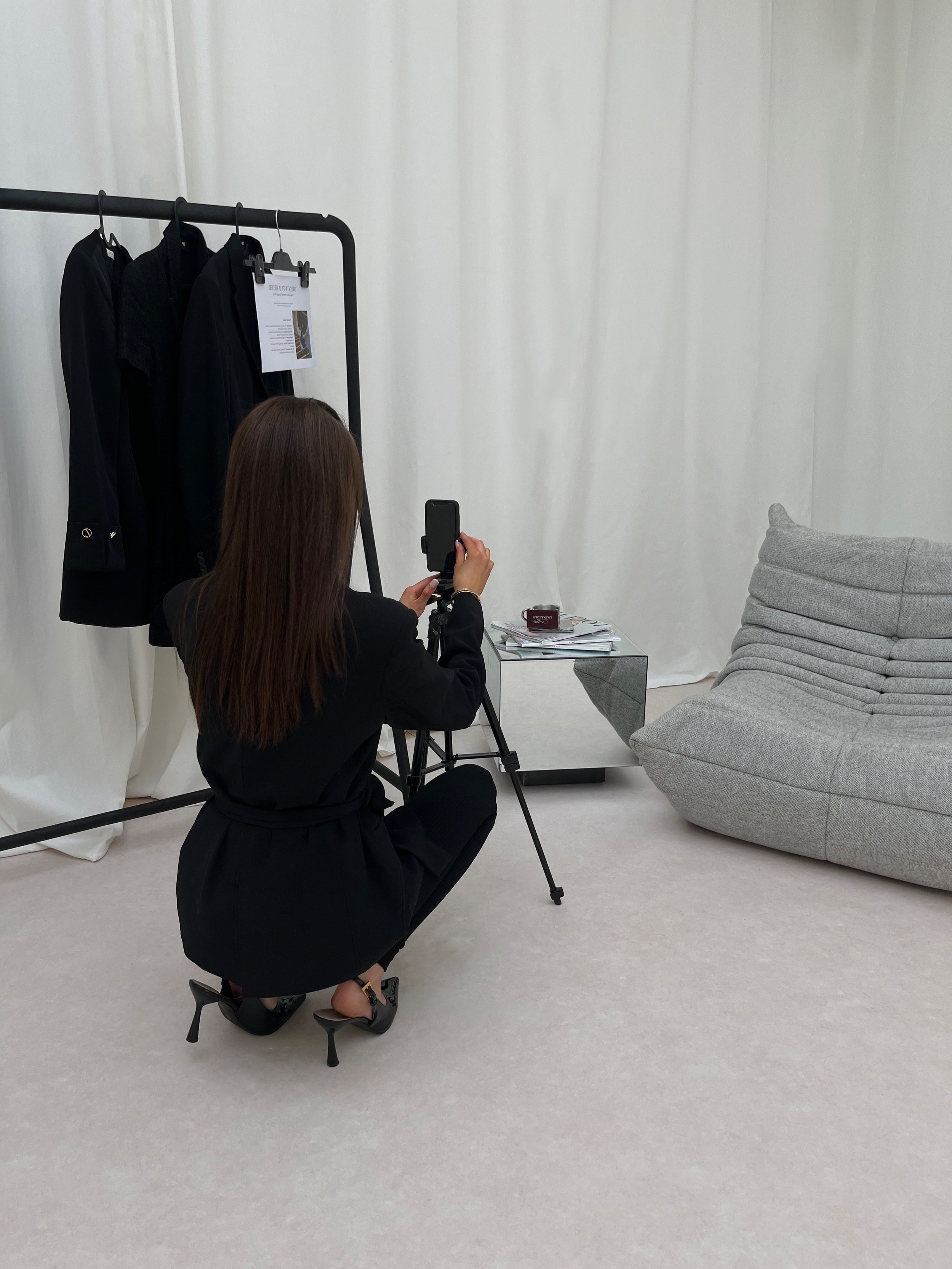 Woman in black clothes crouching and taking a photo of a clothing rack with black blazers or jackets in a studio with white curtains, a gray chair, and a table with papers.