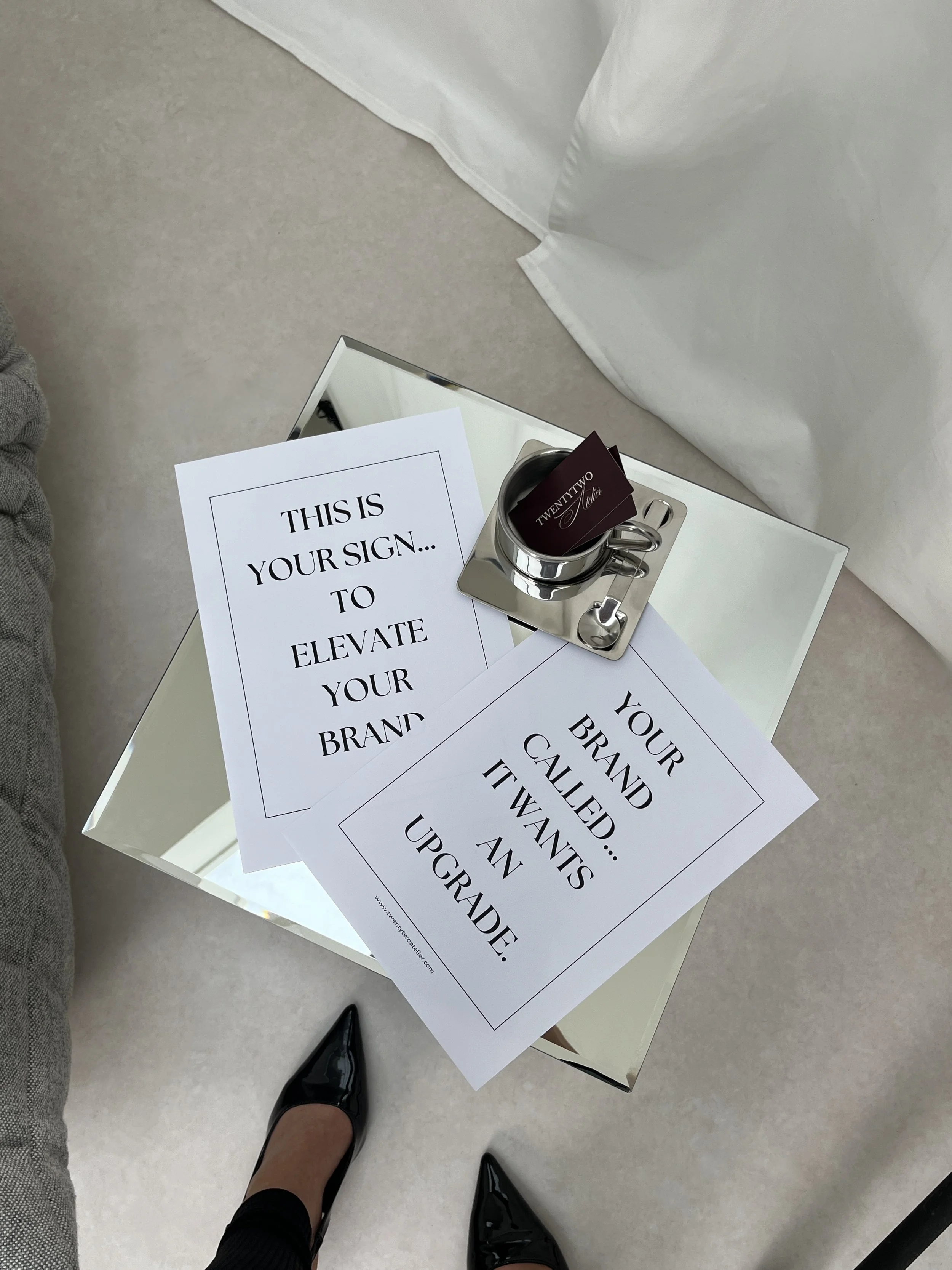 A glass tabletop with two printed signs, a coffee mug, and a pair of black heels. The signs read 'This is your sign to elevate your brand' and 'Your brand called... It wants an upgrade.'