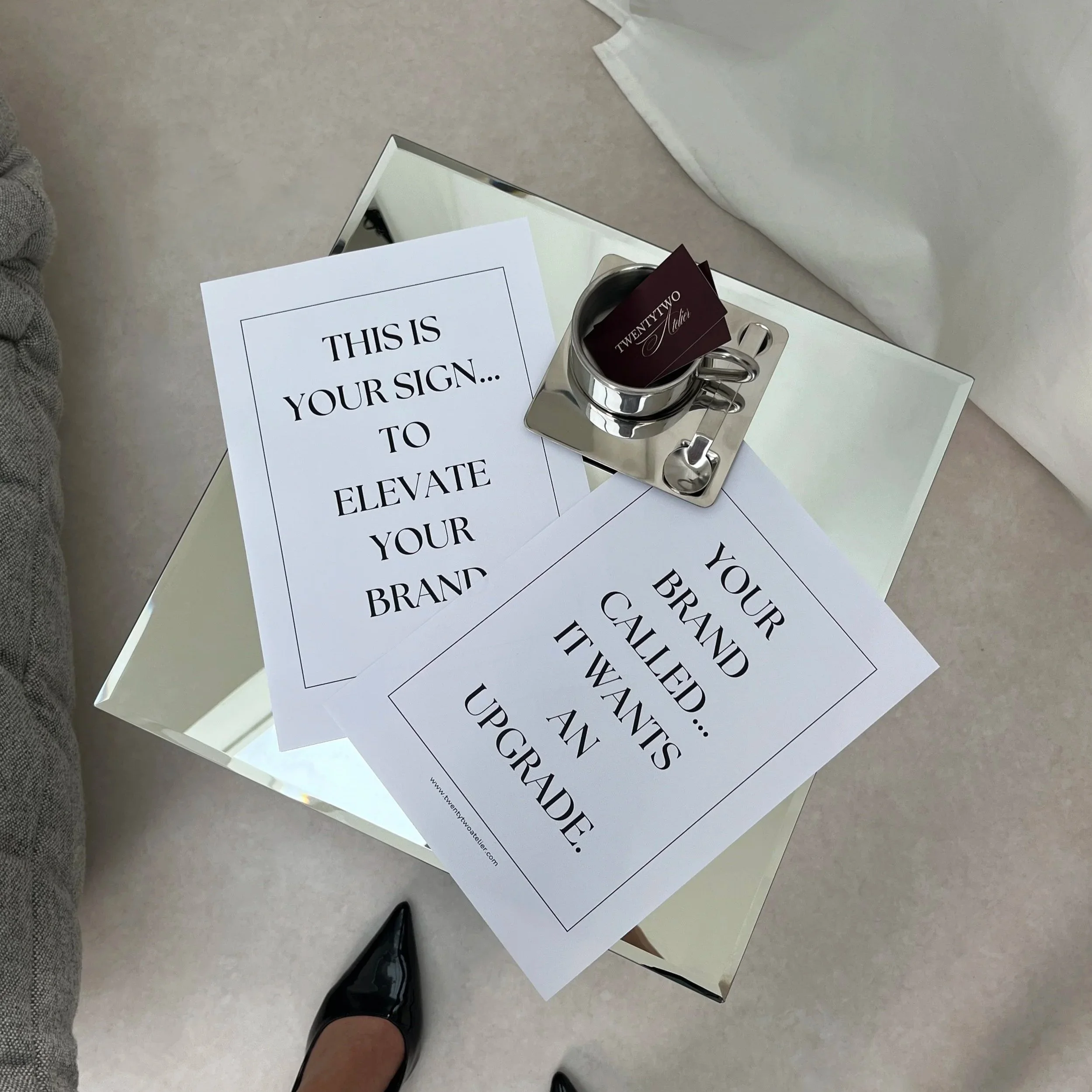 A small side table with two printed signs on it, a silver cup with business cards, and a mirror. One sign says 'This is your sign to elevate your brand' and the other says 'Your brand called... it wants an upgrade.' A person's black high heel shoe is partially visible at the bottom of the image.