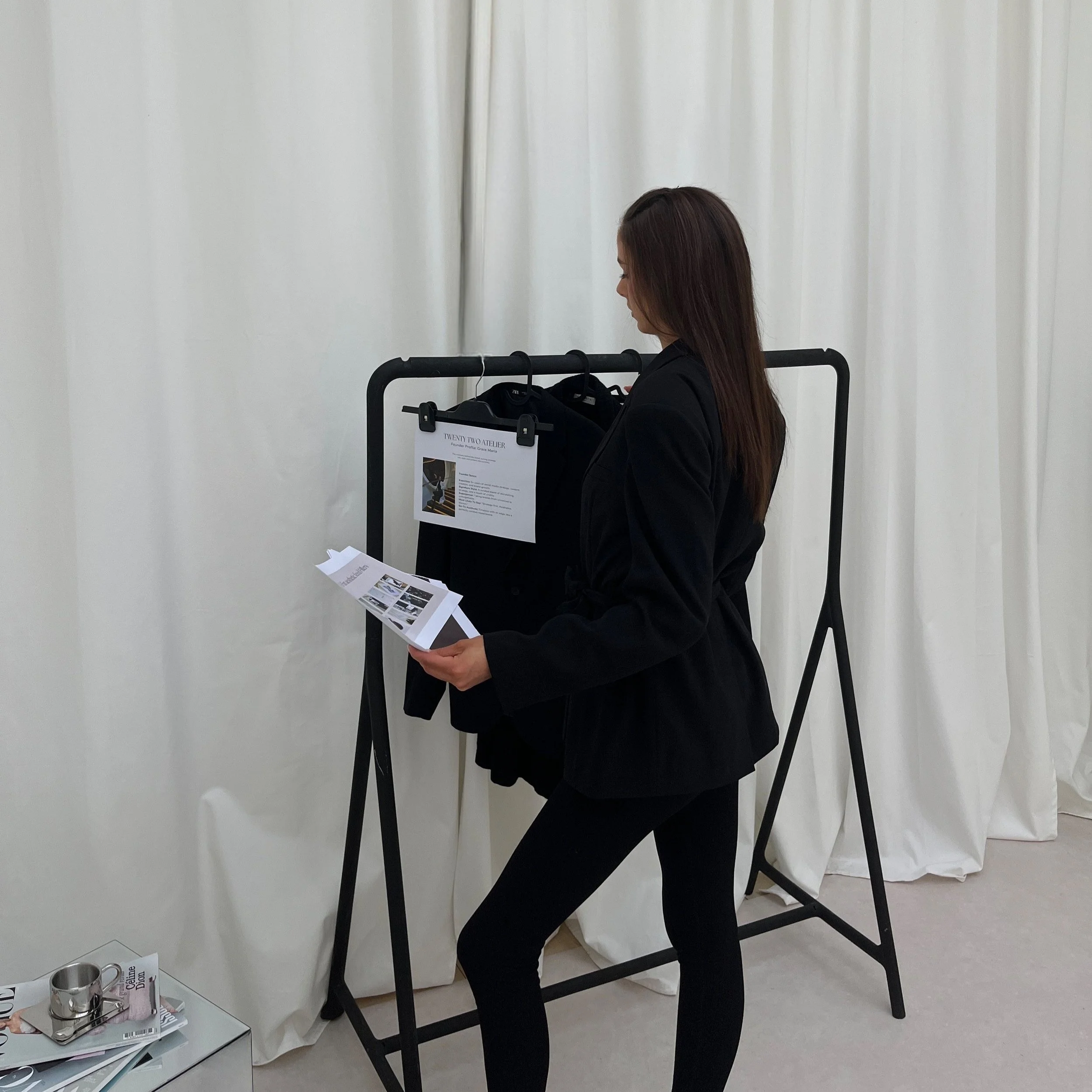 A woman in black clothing looking at a brochure while standing next to a clothing rack with black garments and a hanging paper on it in an indoor setting with white curtains in the background.
