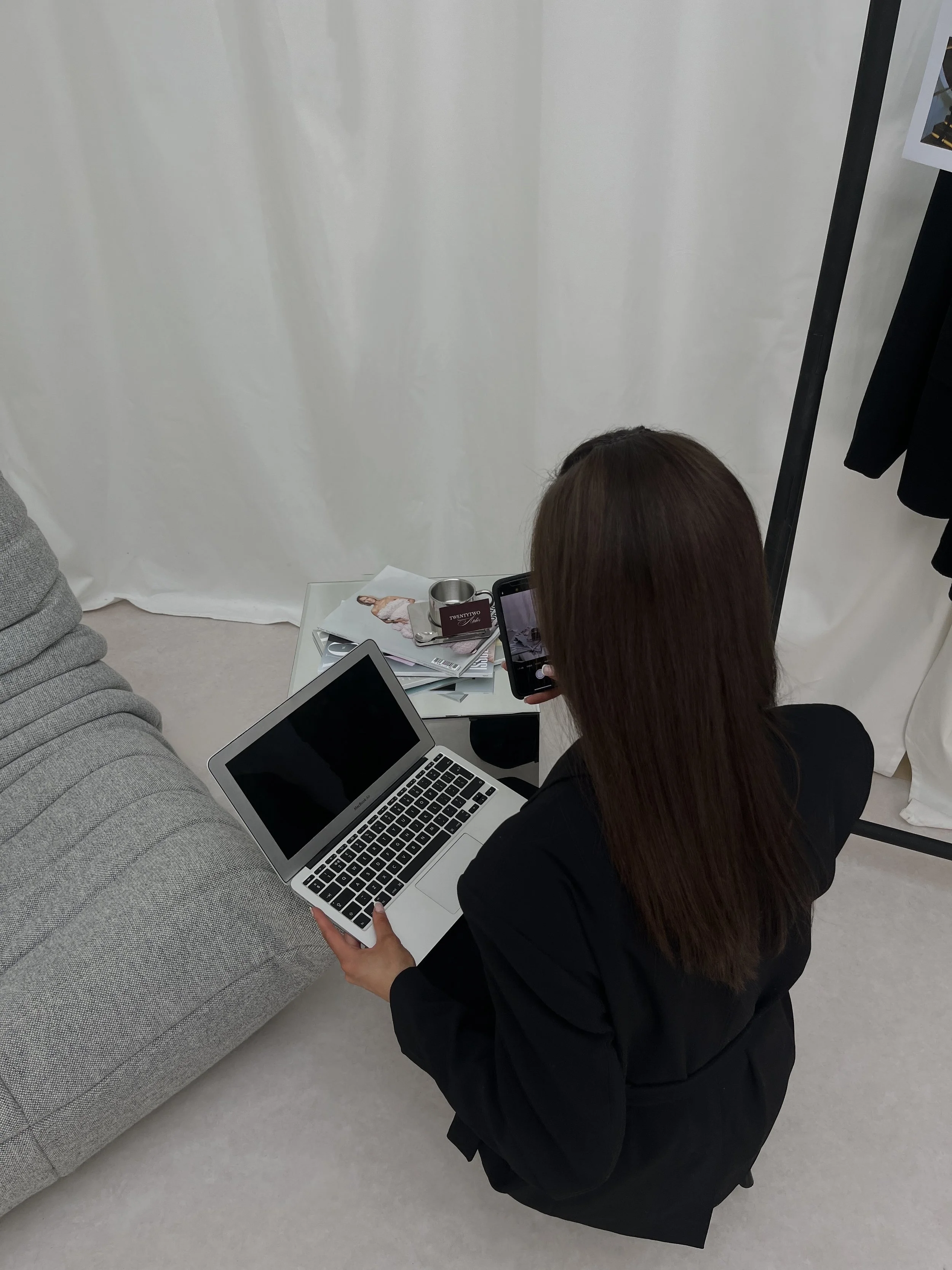 A woman with brown hair in a black blazer is kneeling on the floor, holding a silver MacBook laptop, in a room with white curtains and a display rack. There is a white side table with magazines, a stainless steel mug, and some photos or art prints on top.