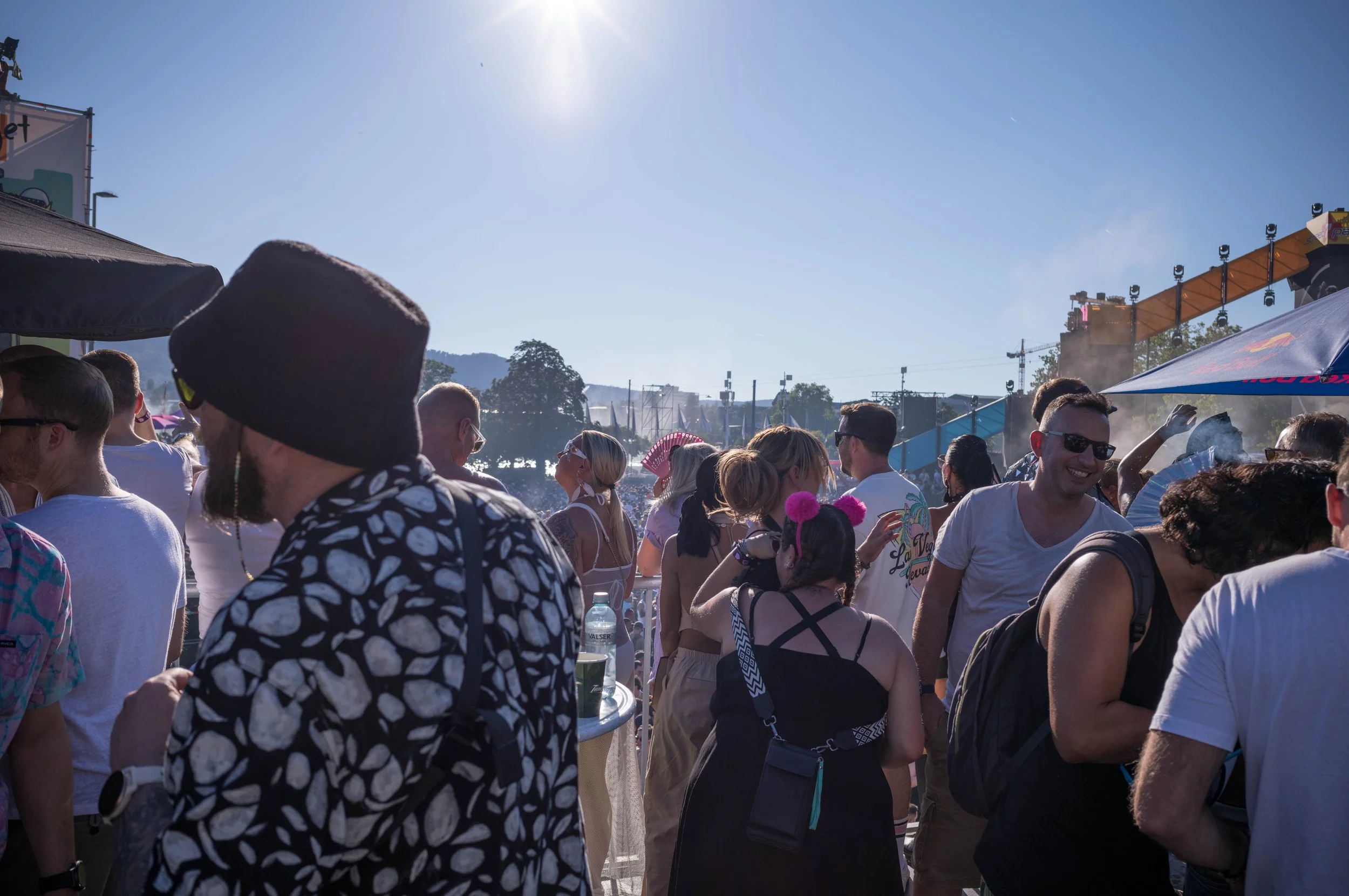 Menschen auf einem Musikfestival im Freien bei Sonne, mit Bühnenaufbau im Hintergrund.