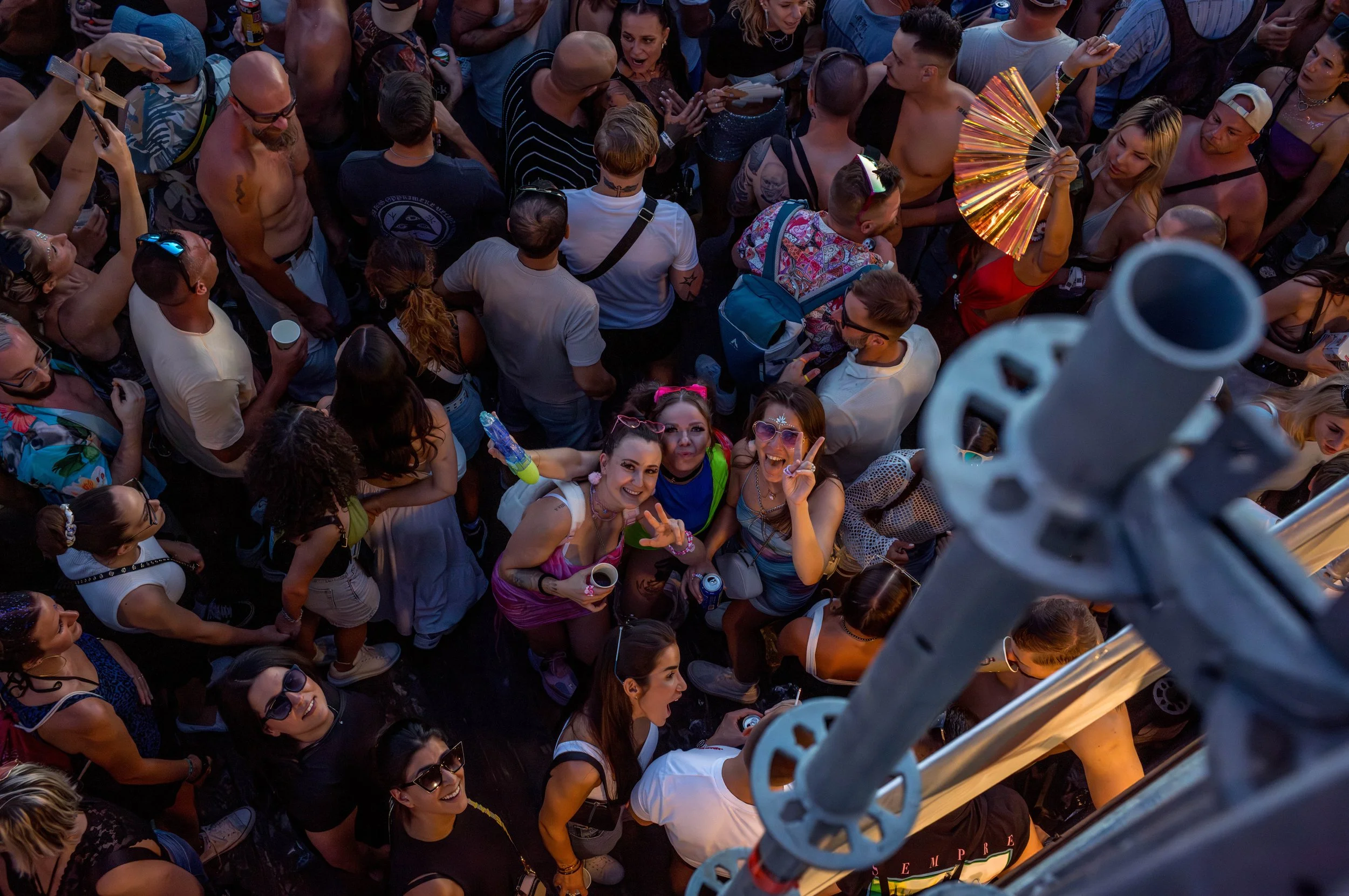Menschen auf einem Festival, einige tanzen, trinken und posieren für die Kamera, während eine Person in der Mitte einen Fächer hält. Die Aufnahme ist von oben.