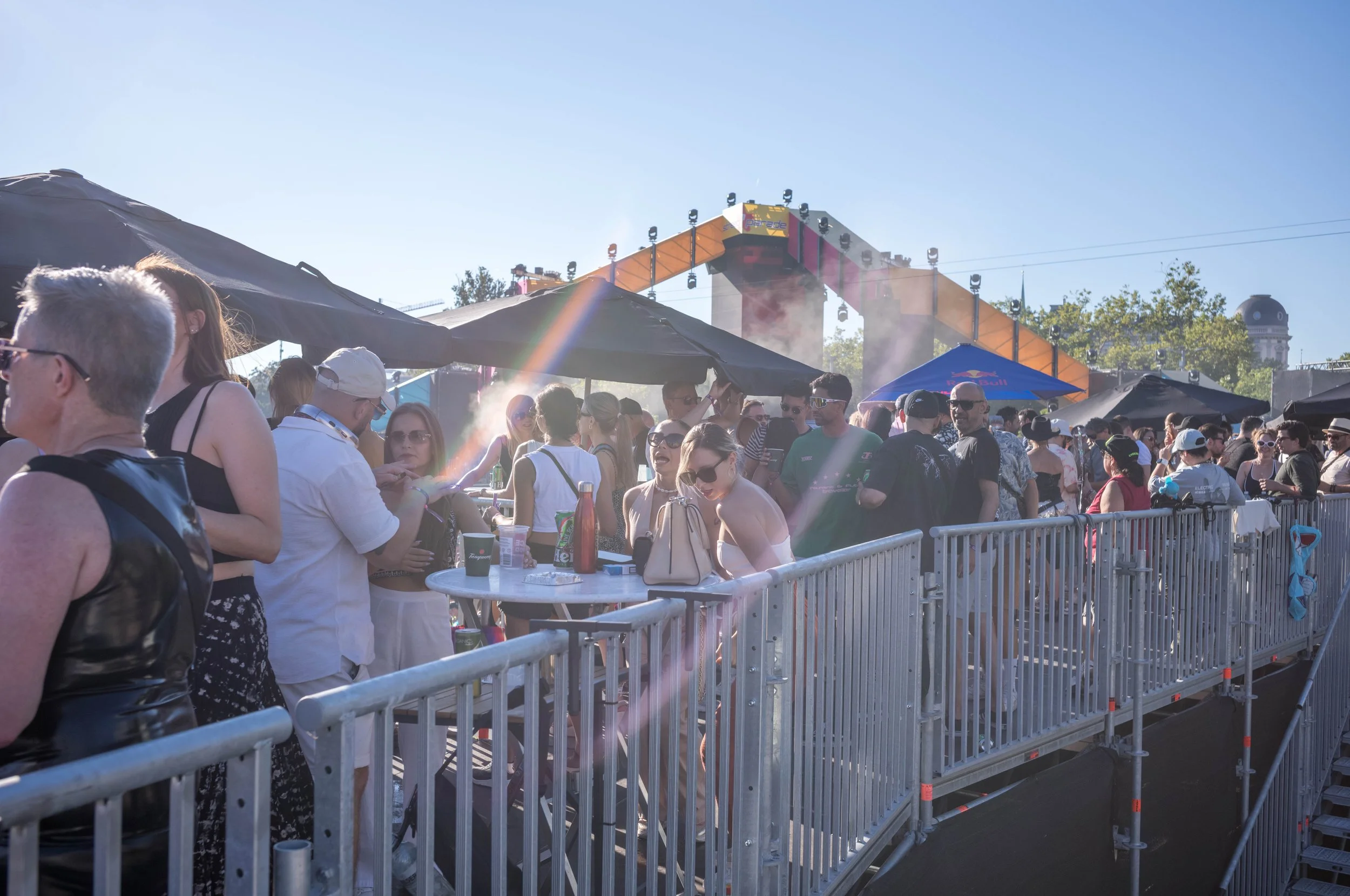 Menschen auf einem Festival vor einer Bühne, einige sitzen an Tischen, andere stehen. Es ist sonnig mit einem klaren Himmel, und es gibt Zelte und Absperrungen.