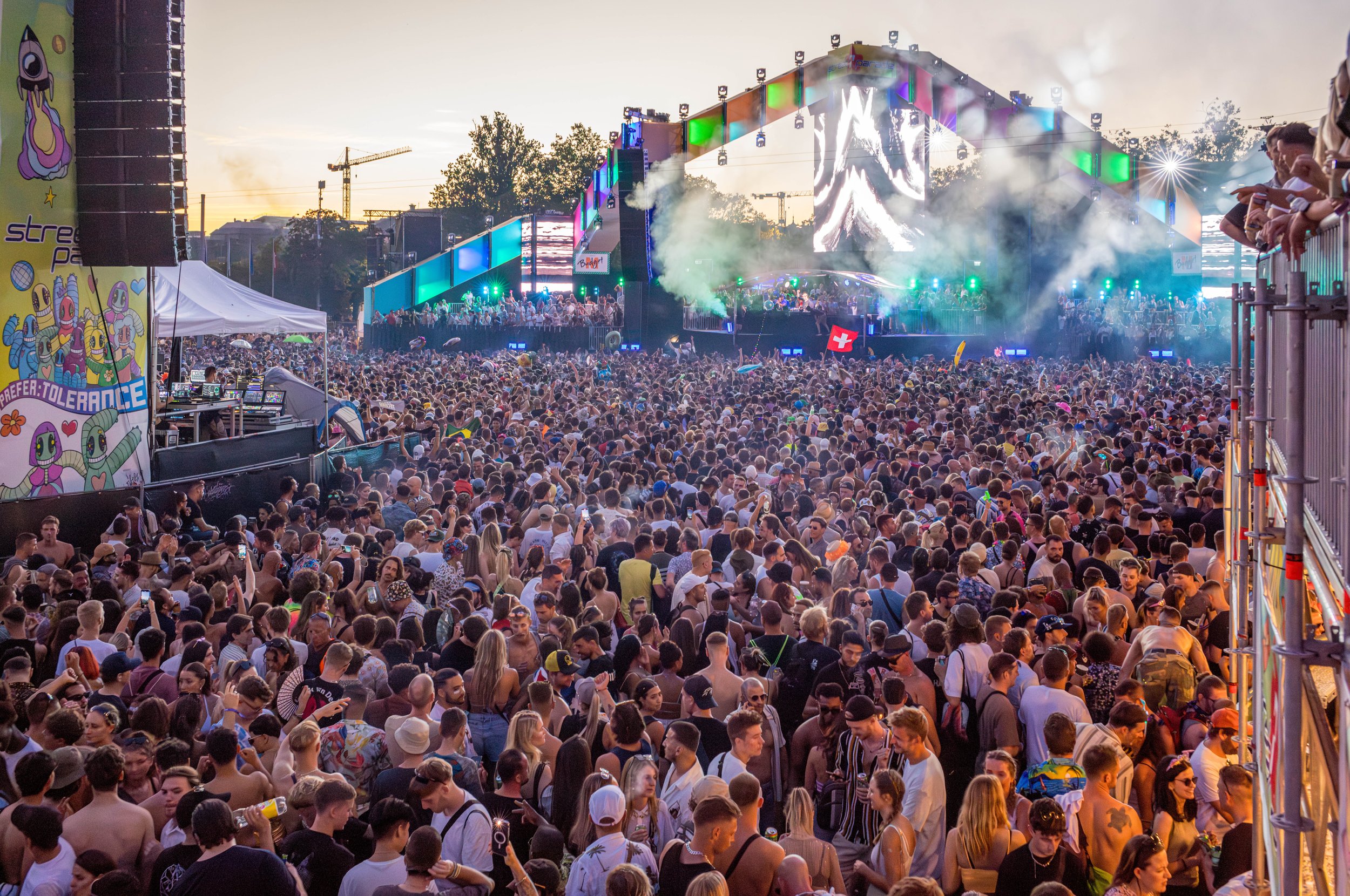 Blick auf eine große Menschenmenge beim Open-Air-Musikfestival, Bühne mit Lichtshow im Hintergrund, während Sonnenuntergang, viele Festivalbesucher in sommerlicher Kleidung, einige mit Hüten, einigen Flaggens.