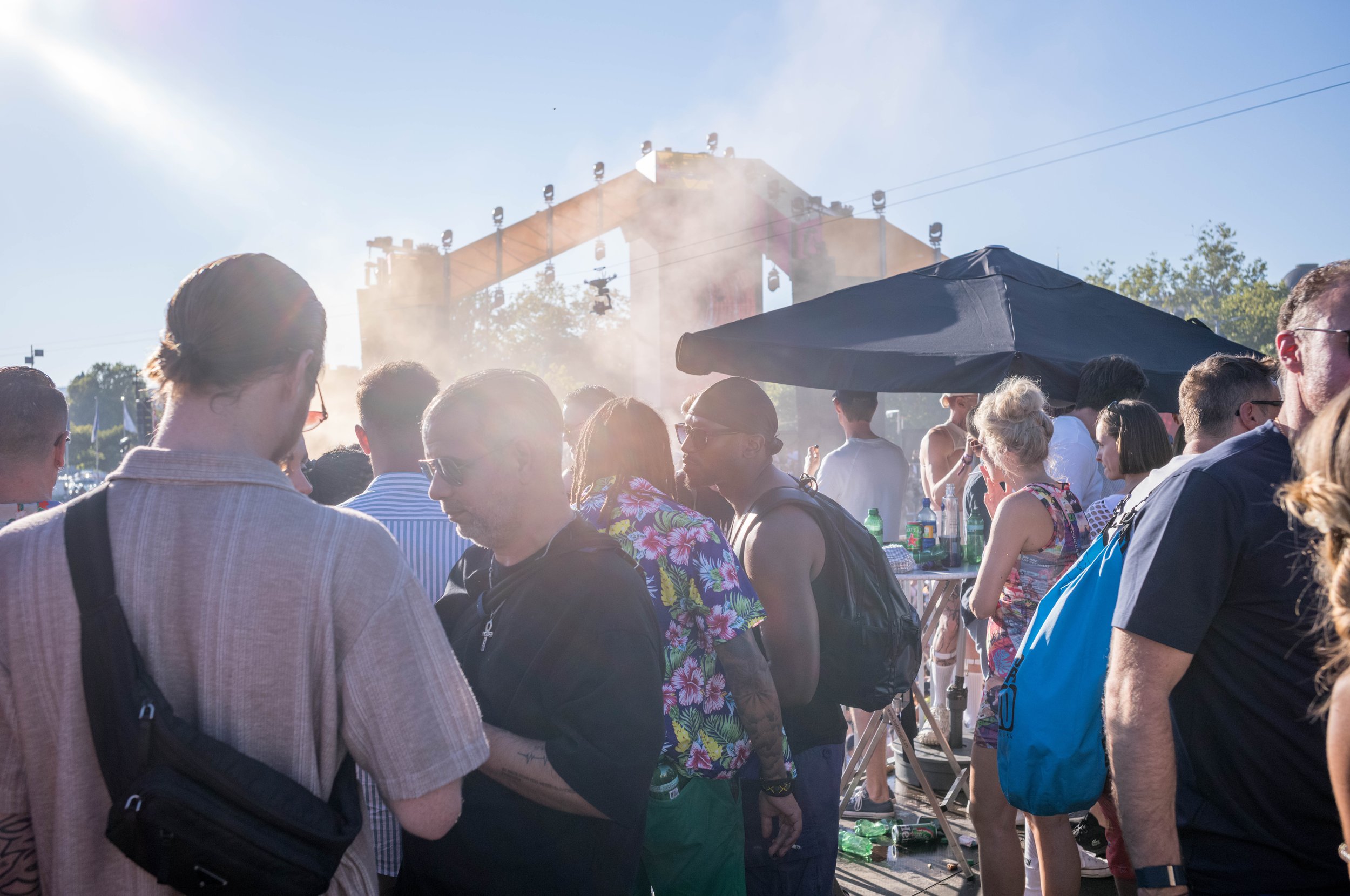Menschen sind auf einem Festival versammelt, mit einer Bühne im Hintergrund und einem großen schwarzen Sonnenschirm, die Sonne scheint, es gibt Getränke auf Tischen.