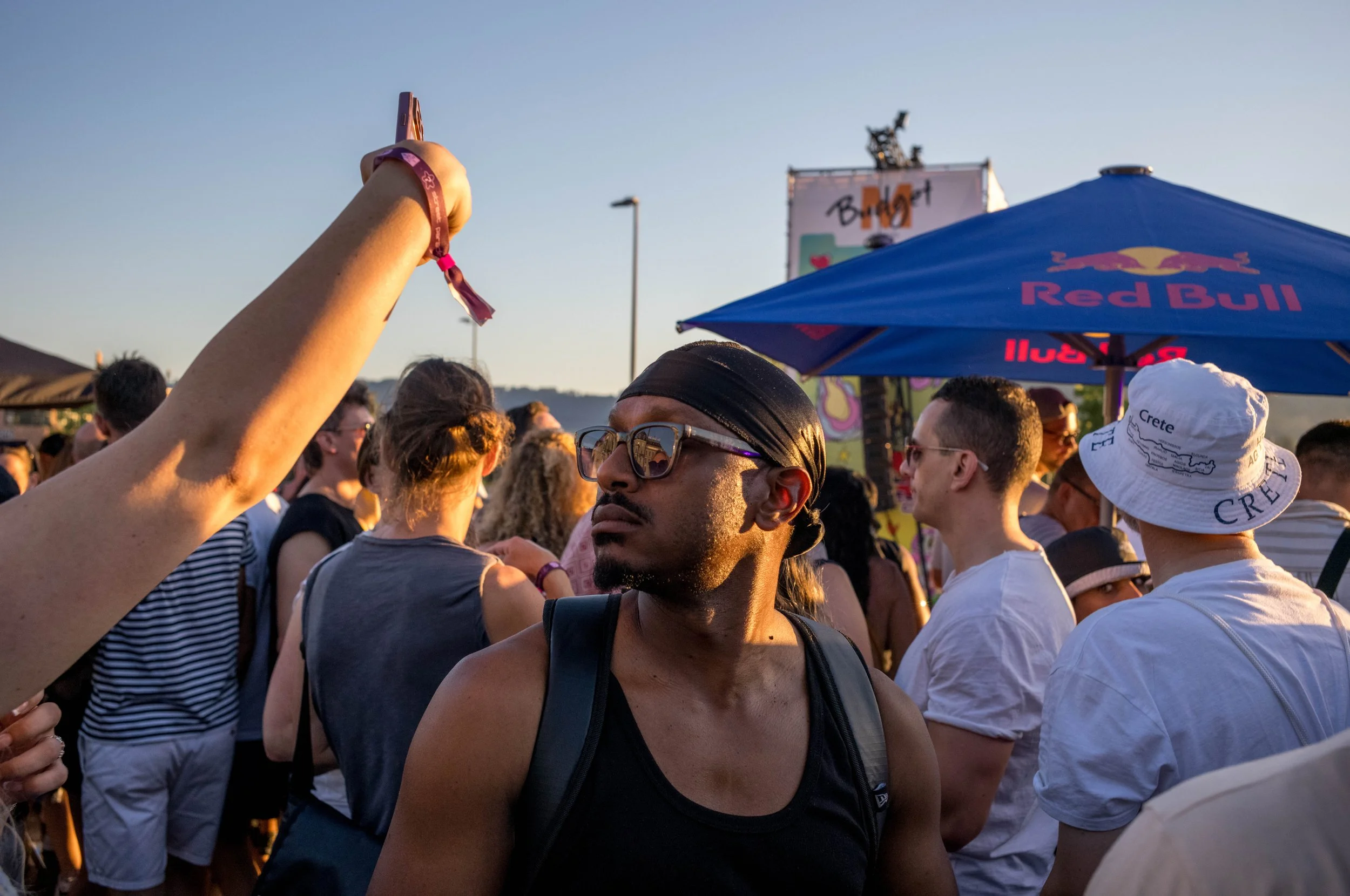 Menschenmenge auf einem Musikfestival im Freien bei Sonnenuntergang, im Vordergrund ein Mann mit Sonnenbrille, schwarz Bandana und schwarzem Tanktop.