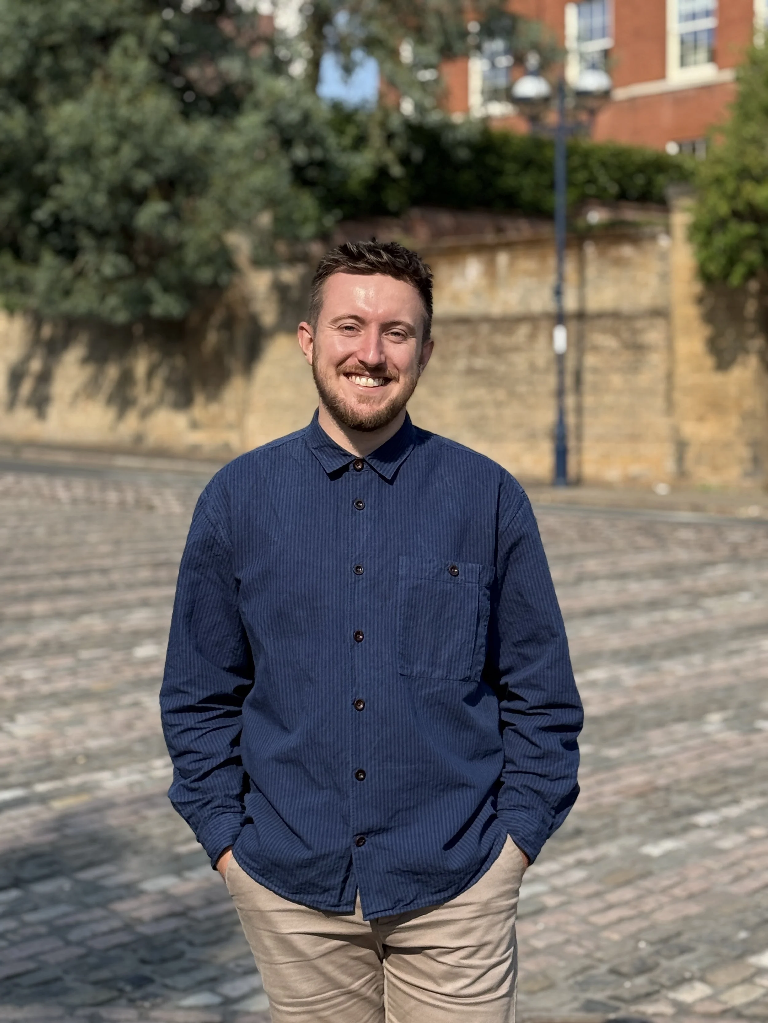 A smiling young man with short dark hair and a beard, wearing a navy blue button-up shirt and beige pants, standing outdoors on a cobblestone street with a brick wall, trees, and a lamppost in the background.