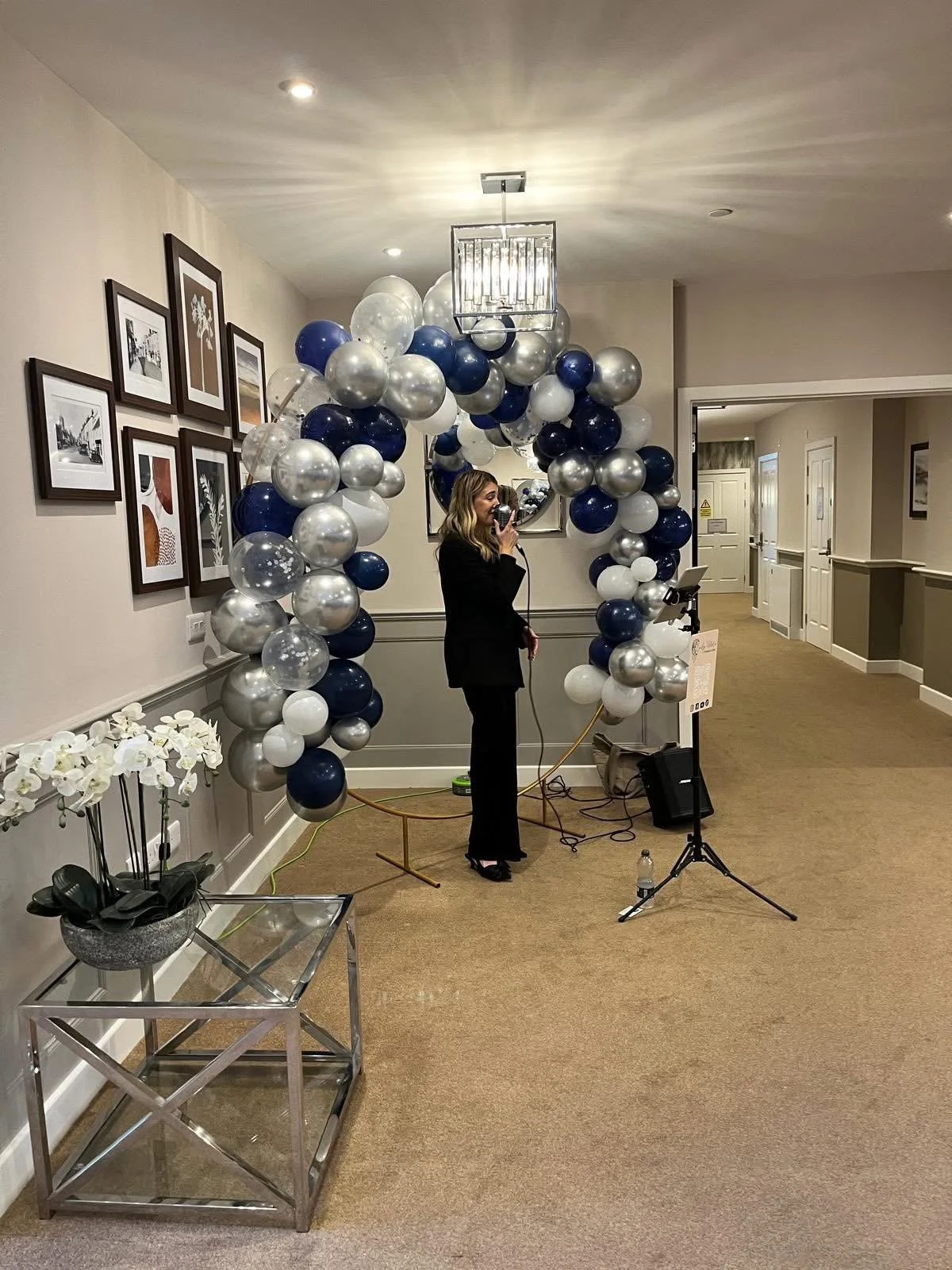 Woman in black suit taking a photo in front of a balloon arch with white, navy blue, and silver balloons at an indoor event.