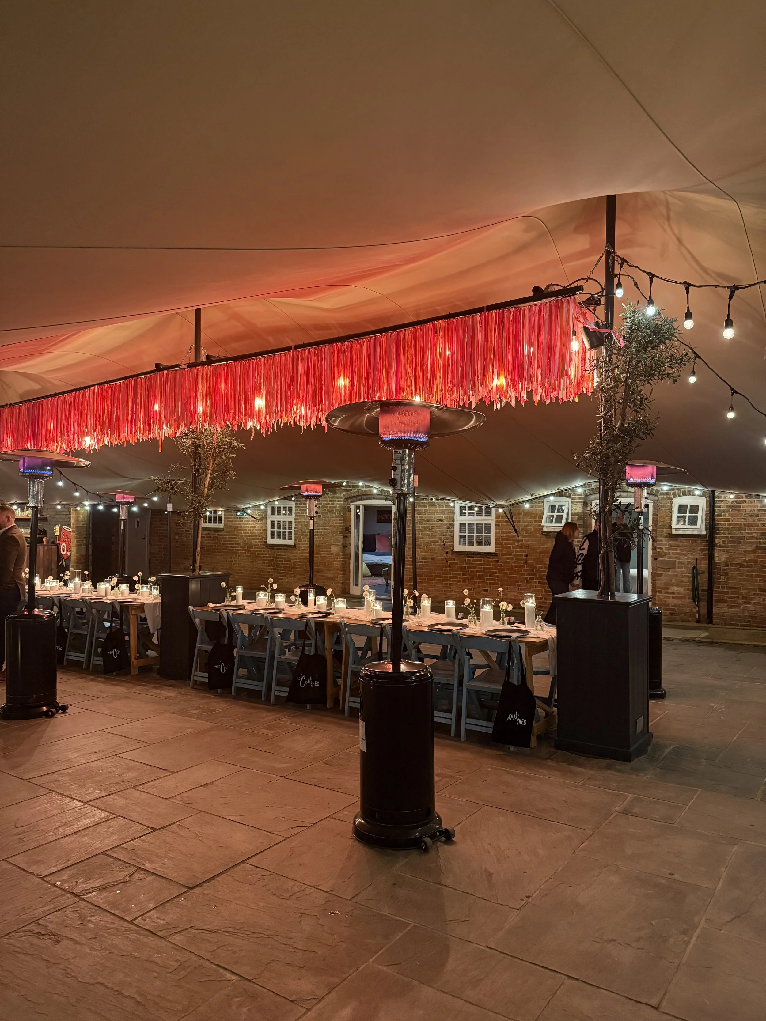 Indoor event space decorated with string lights, palm trees in planters, a long dining table set with candles, and a red tassel fringe decoration hanging from the ceiling.
